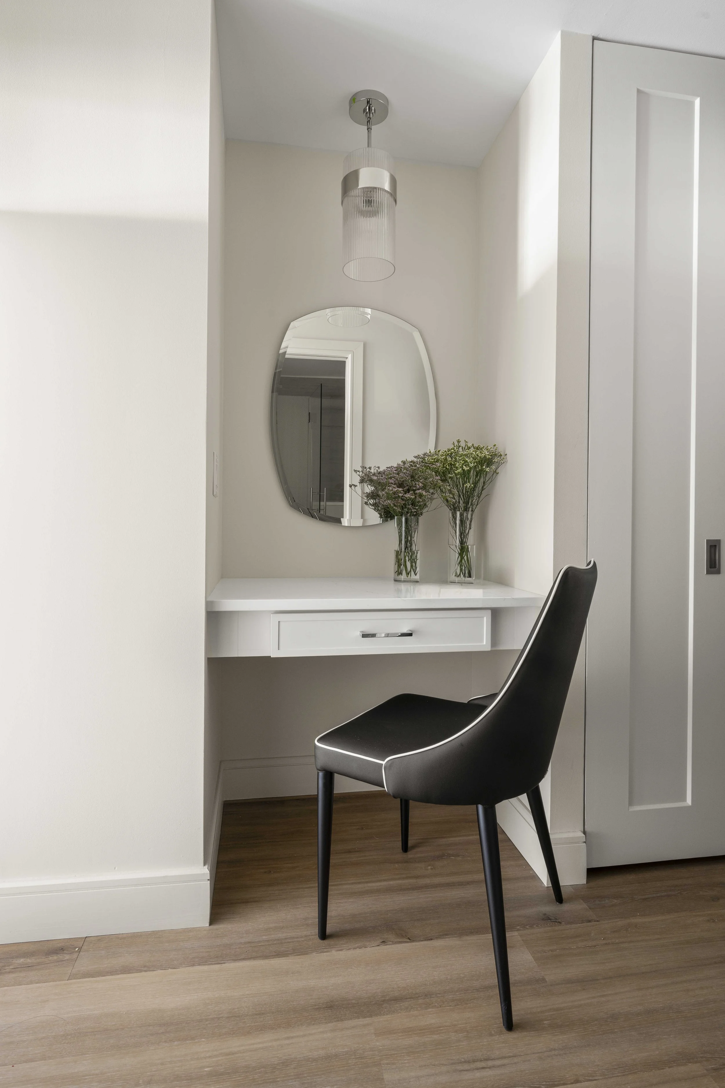 A minimalist built-in vanity nook featuring a white desk with a single drawer, an asymmetrical frameless mirror, and a sleek black chair with white piping. A fluted glass pendant light hangs overhead.