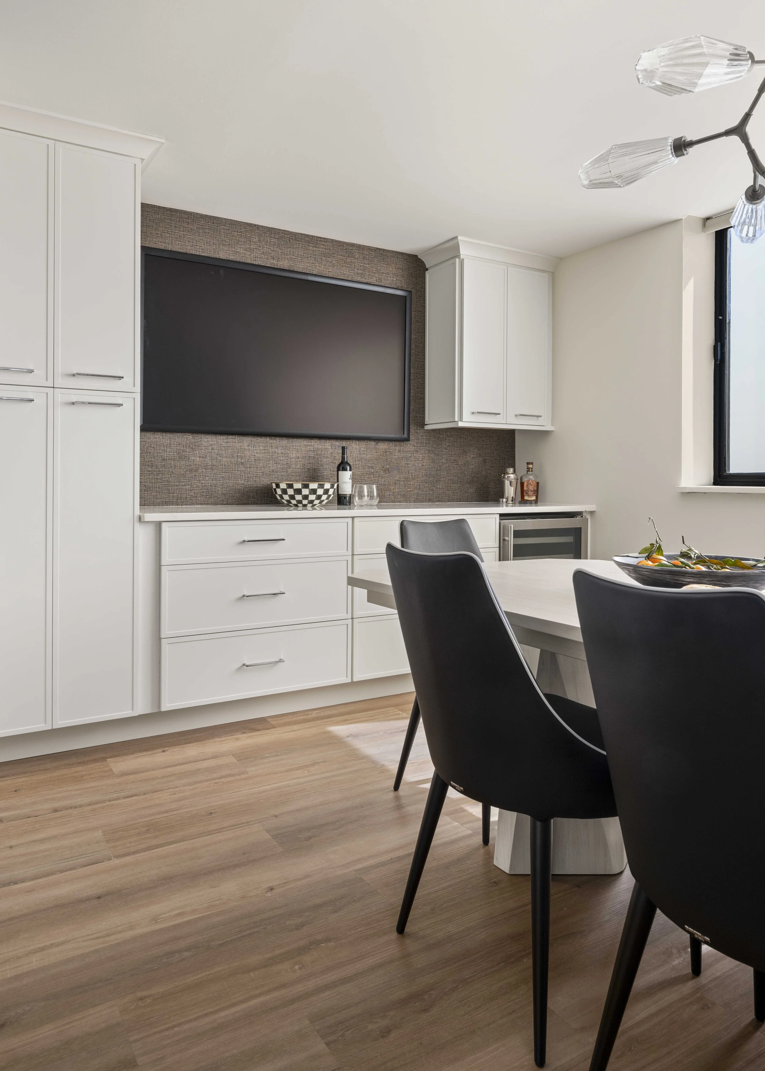 A close-up of a kitchen buffet area with white drawers, a wine cooler, and a large black framed screen mounted on a textured brown wallpaper backsplash.