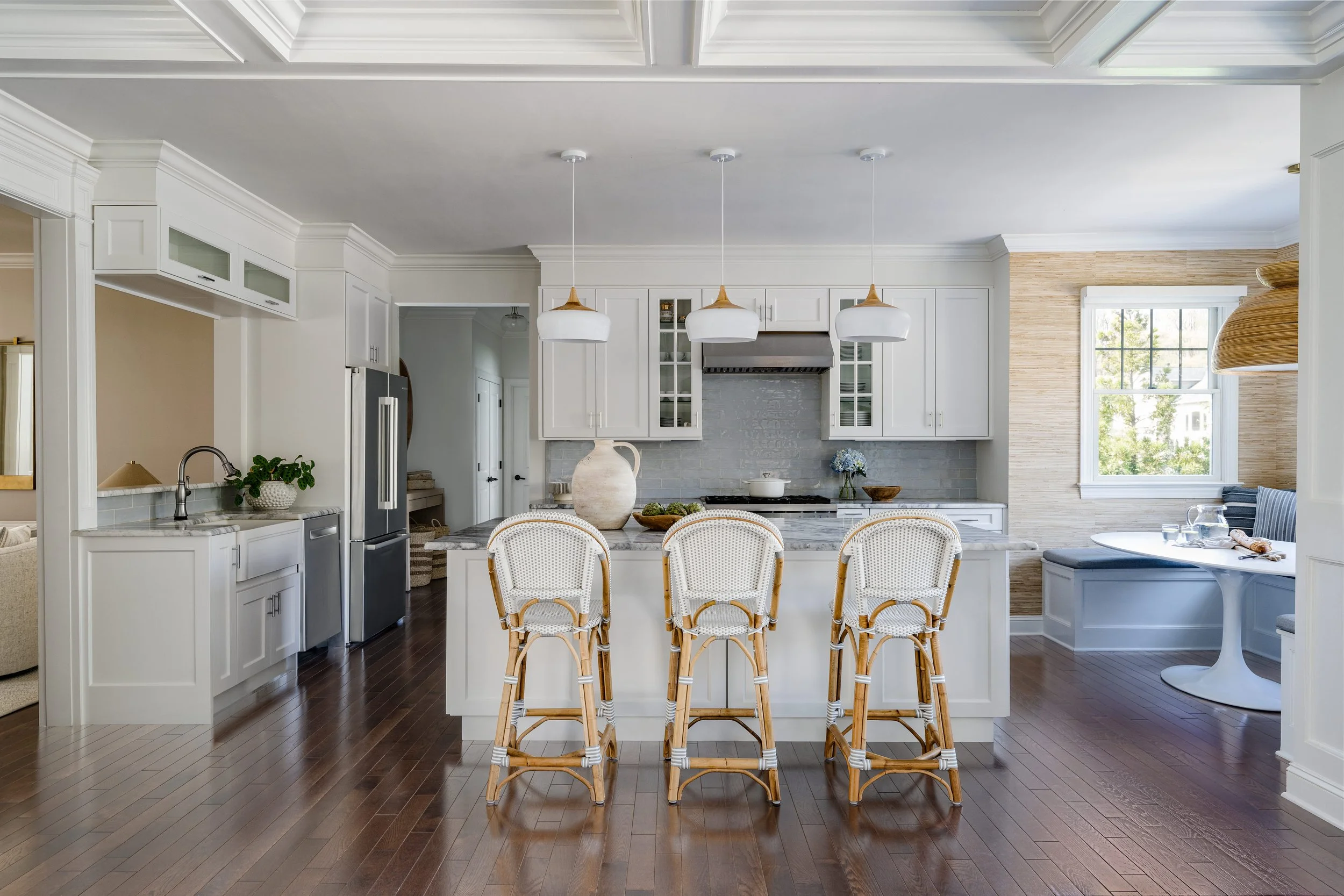 Bright open-concept kitchen with white shaker cabinetry, coffered ceiling, three French bistro-style rattan counter stools at marble waterfall island, brass and white pendant lights, gray subway tile backsplash, stainless appliances, view into breakf