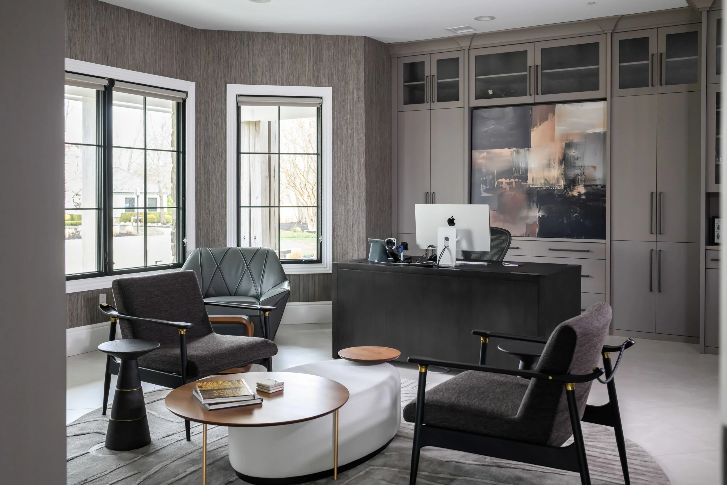 Contemporary home office with black desk, gray built-in cabinetry with glass-front uppers, abstract artwork, curved white ottoman, gray and wood accent chairs with brass details, and black-framed windows with textured wallcovering.