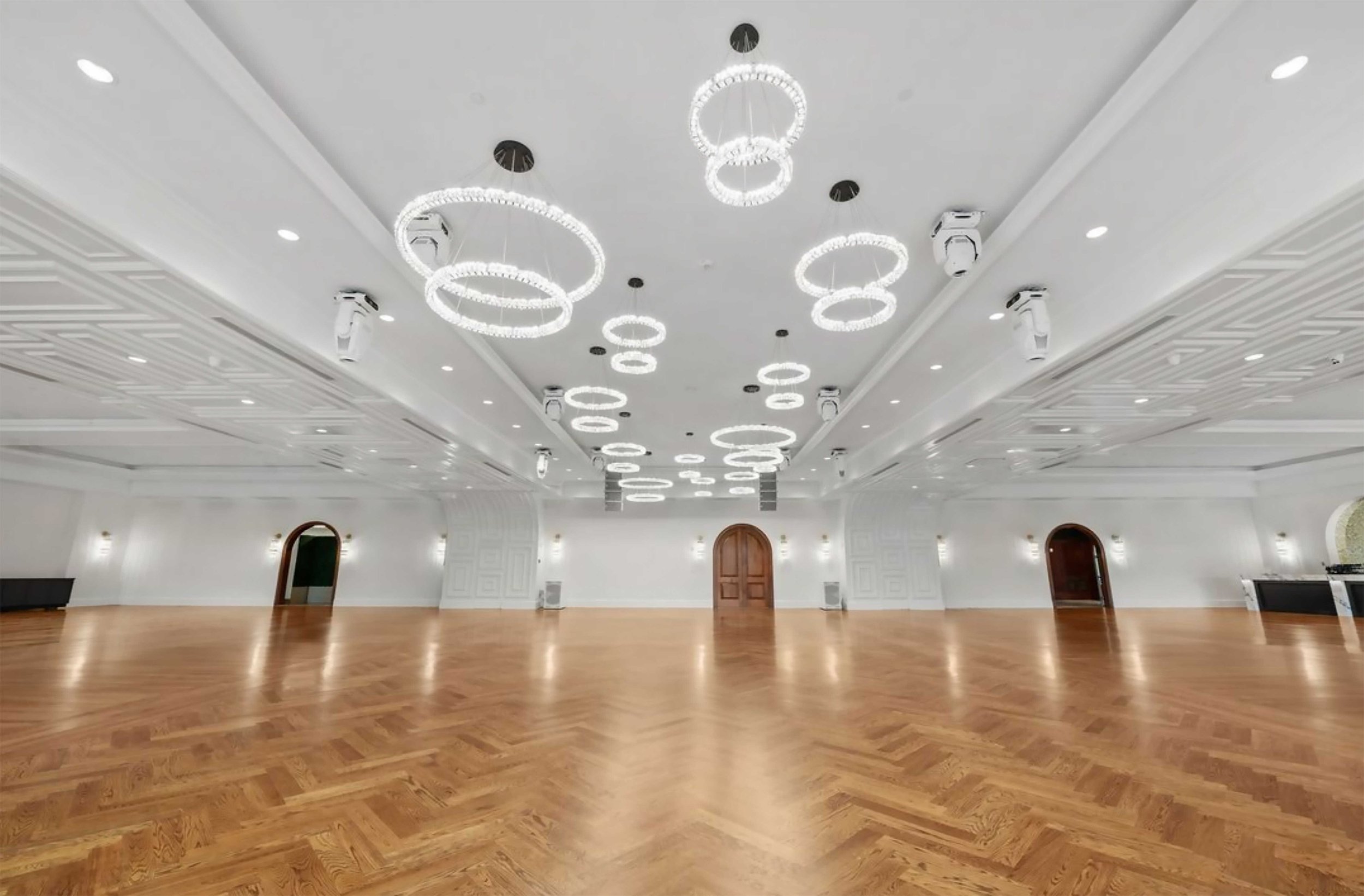 An expansive ballroom with light oak herringbone floors and a ceiling decorated with numerous modern, glowing circular ring chandeliers.