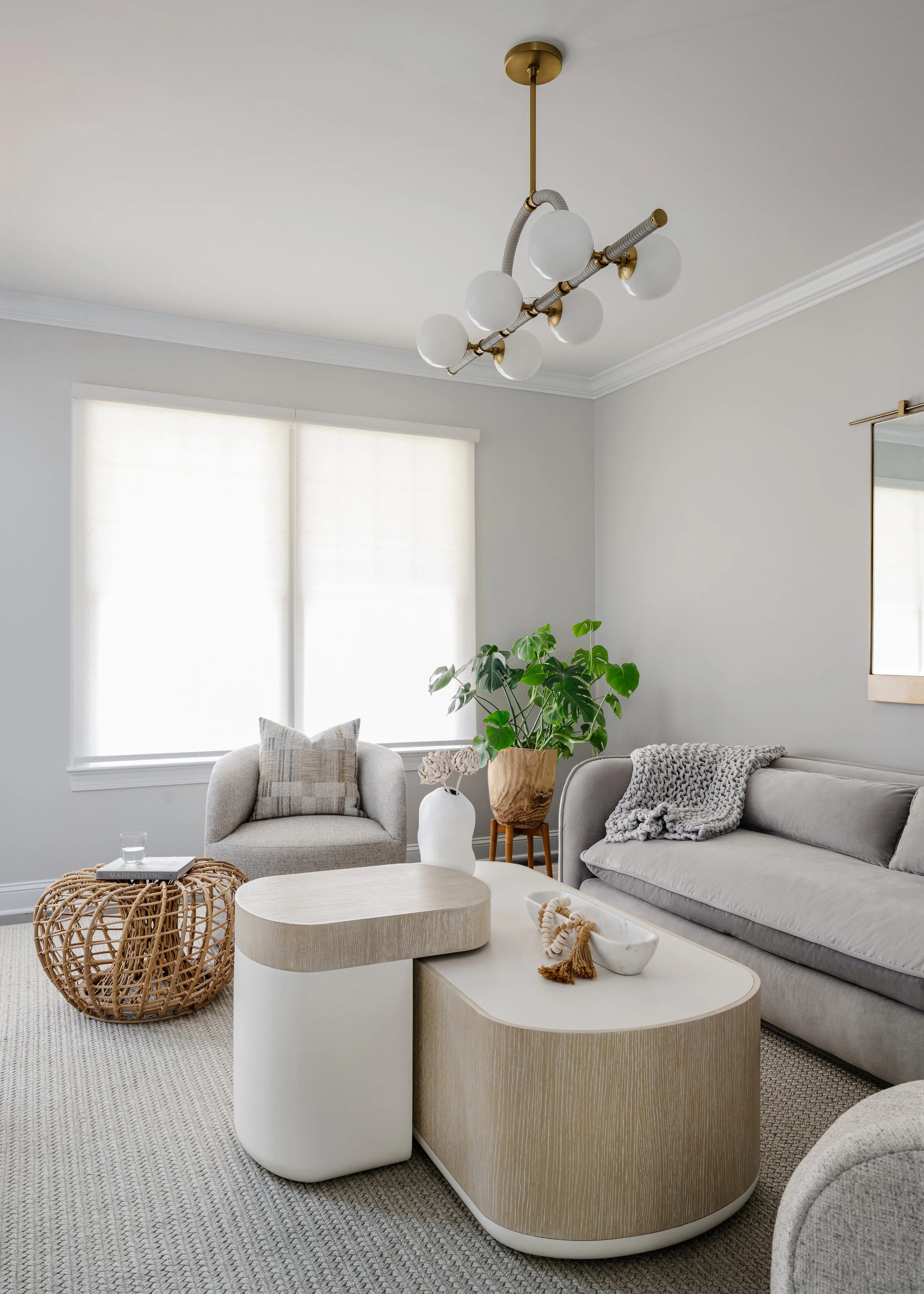 Detail of living room showing modern brass chandelier with frosted glass globe shades on asymmetrical arms, gray sofa, nested cream and wood coffee tables with white ceramic vase and natural rope accent, woven basket ottoman, and plant stand with gre