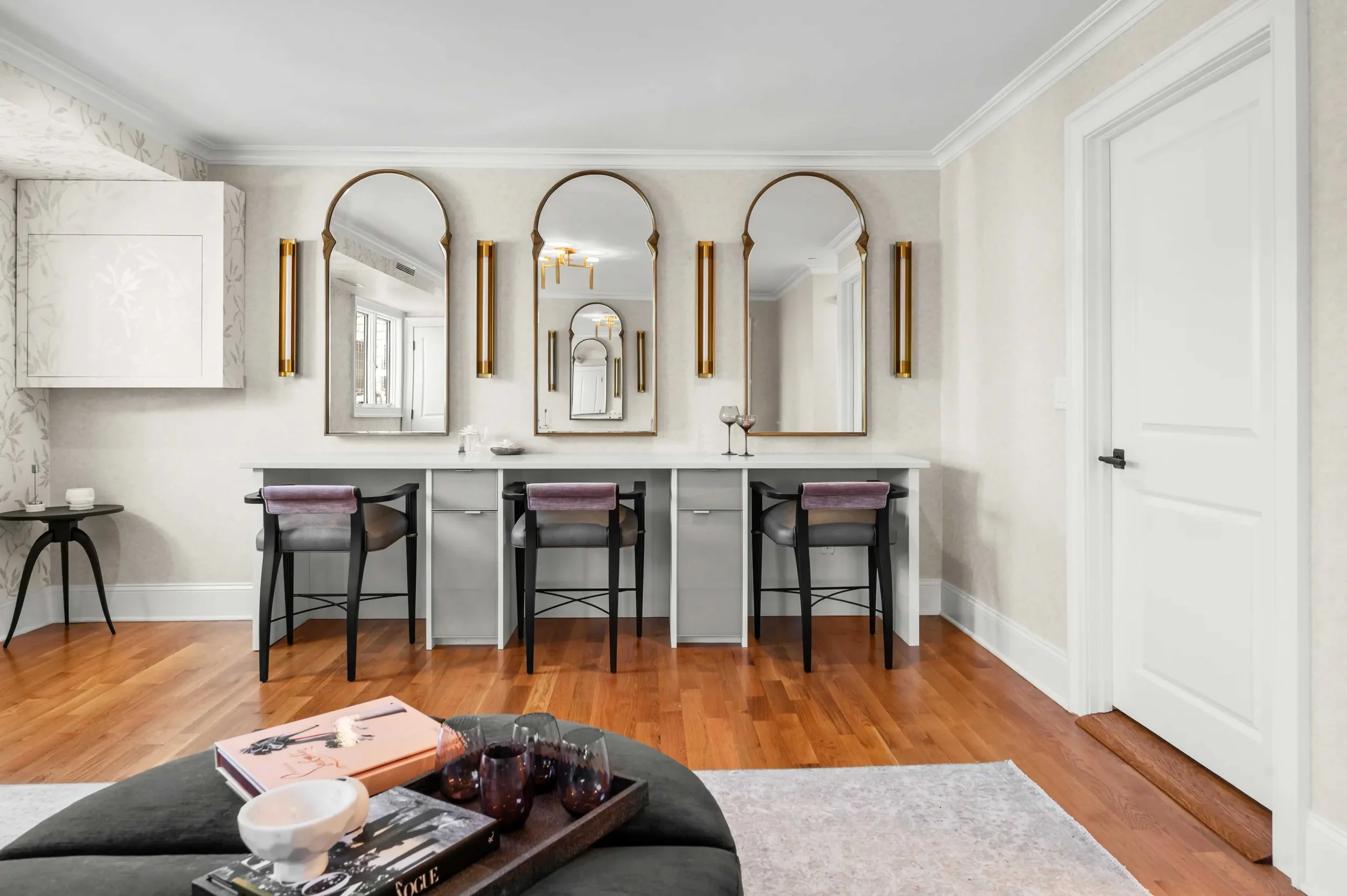A spacious and elegant dressing room featuring a long white vanity with three built-in stations. Each station includes a grey and purple cushioned chair, a tall arched gold-framed mirror, and vertical brass wall sconces. The room is finished with war