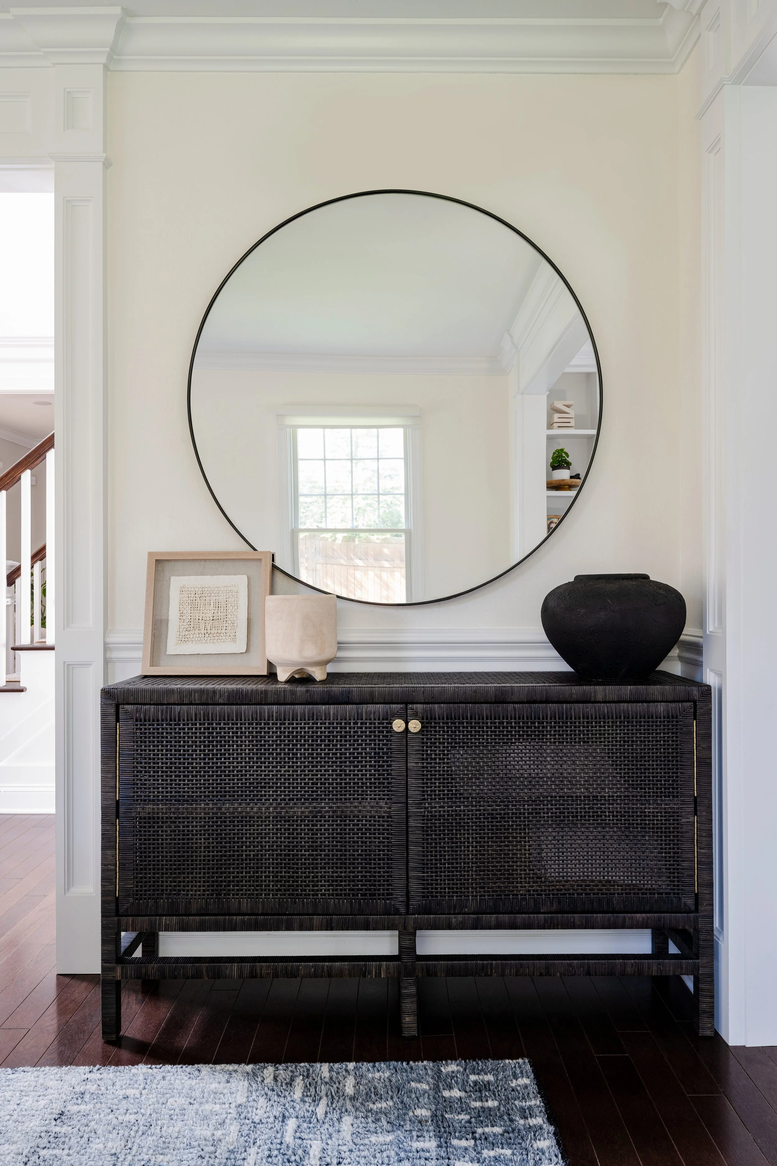 Traditional entryway with oversized round black-framed mirror above textured black raffia console with brass knobs, styled with framed neutral artwork, cream ceramic vessel, and black pottery, dark wood floors with blue-gray area rug, white wainscoti