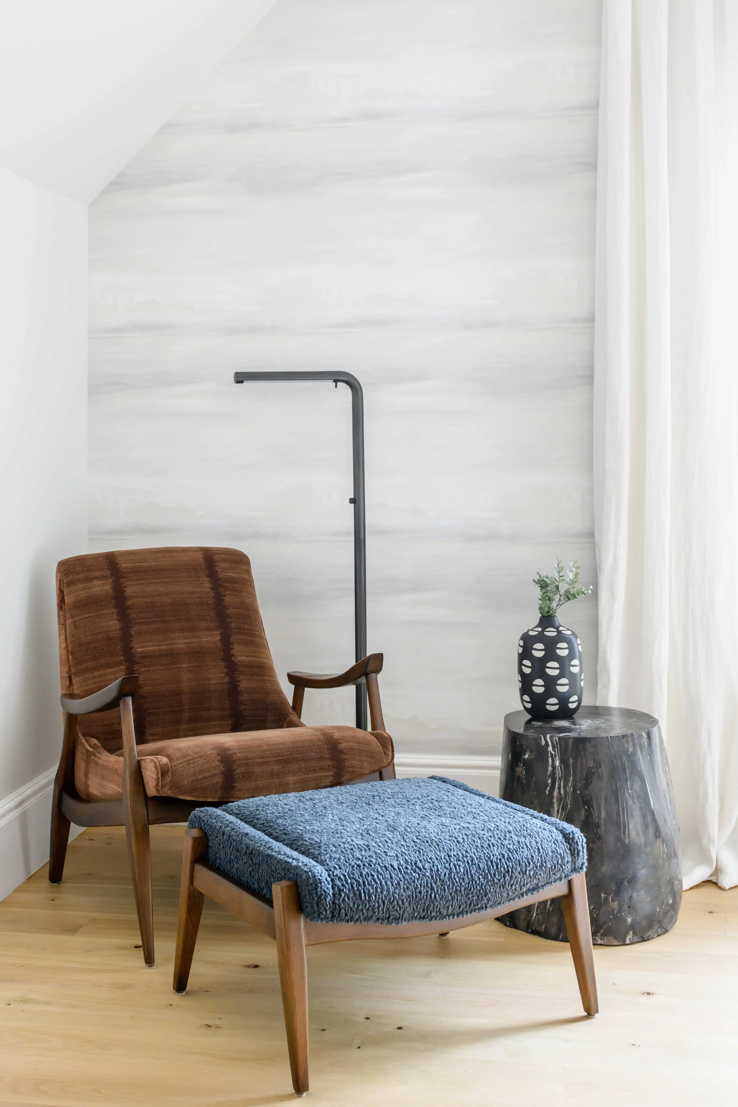 Cozy reading nook with mid-century modern walnut lounge chair featuring rust velvet striped upholstery, blue sherpa ottoman, black adjustable floor lamp, black marbled side table holding polka-dot vase with white flowers, and soft textured wallcoveri