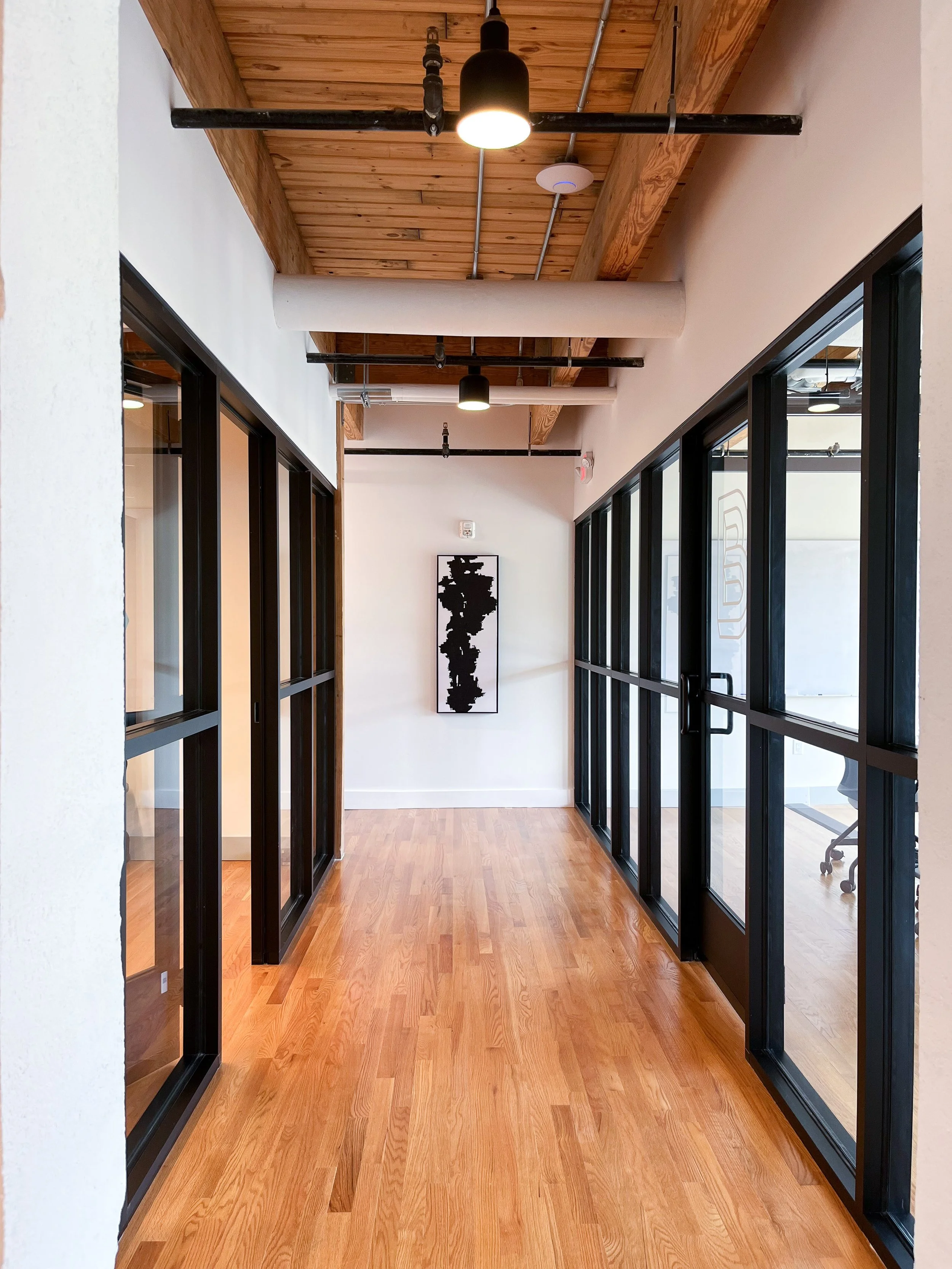 Modern office corridor with natural wood plank ceiling featuring exposed black beams, recessed ceiling panels, white walls, light wood flooring, black-framed glass office partitions on both sides, black pendant lights, and vertical abstract artwork a