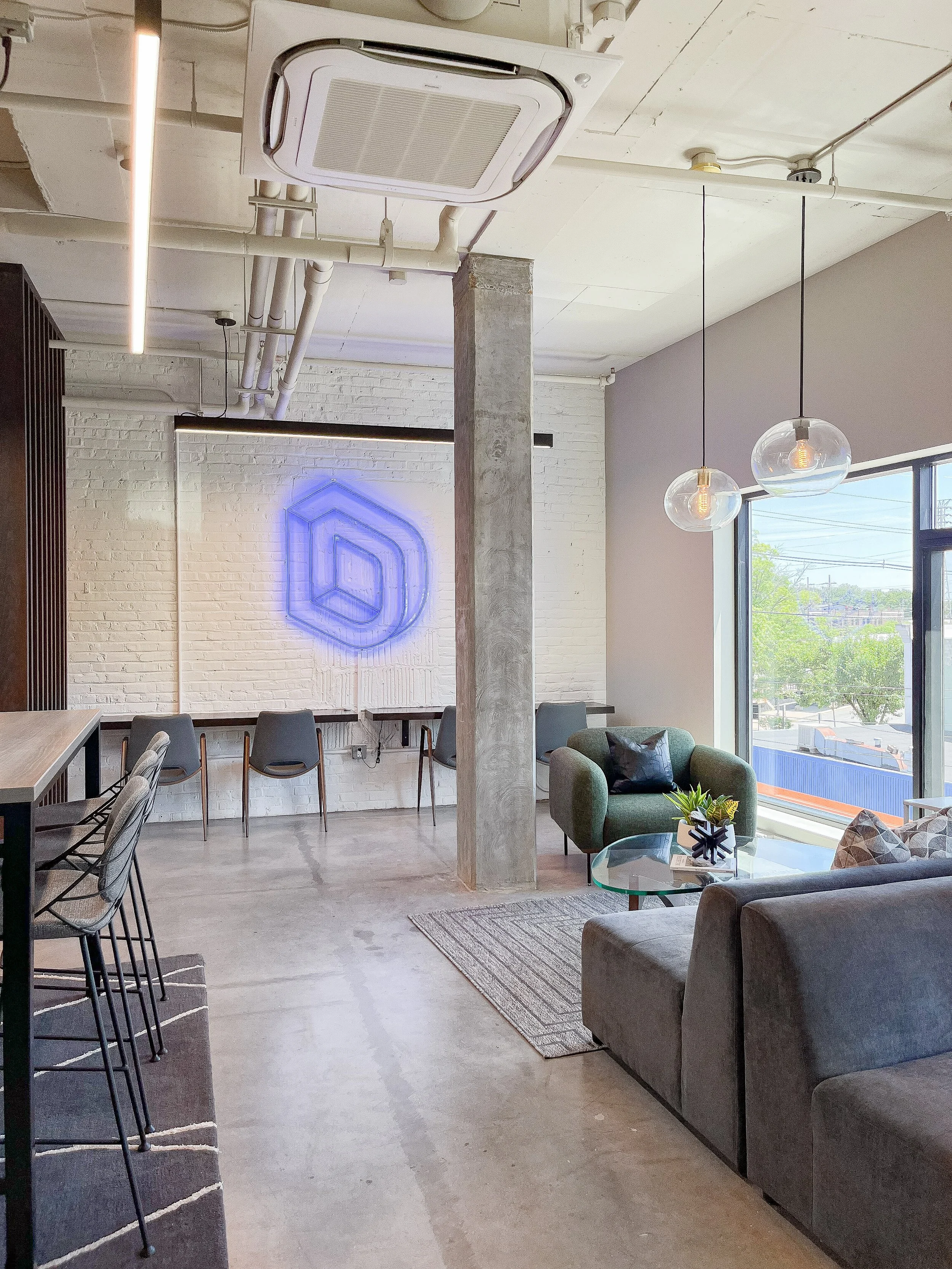 Industrial office space with polished concrete floors, exposed white brick walls, concrete structural columns, gray sofas with glass coffee table, emerald green velvet chairs, blue geometric LED artwork mounted on wall, wire-frame modern chairs at hi