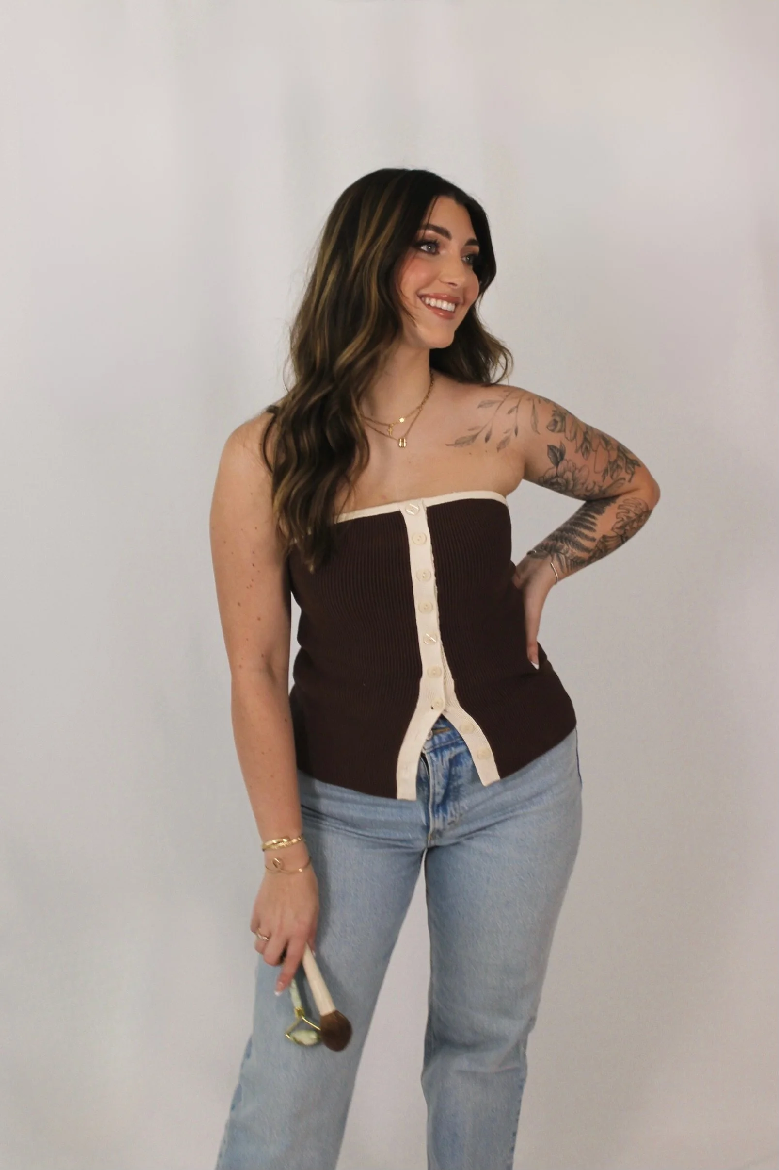 A young woman with long brown hair, smiling, wearing a strapless brown and cream button-up top, light blue jeans, and holding a makeup brush, standing against a plain white background.