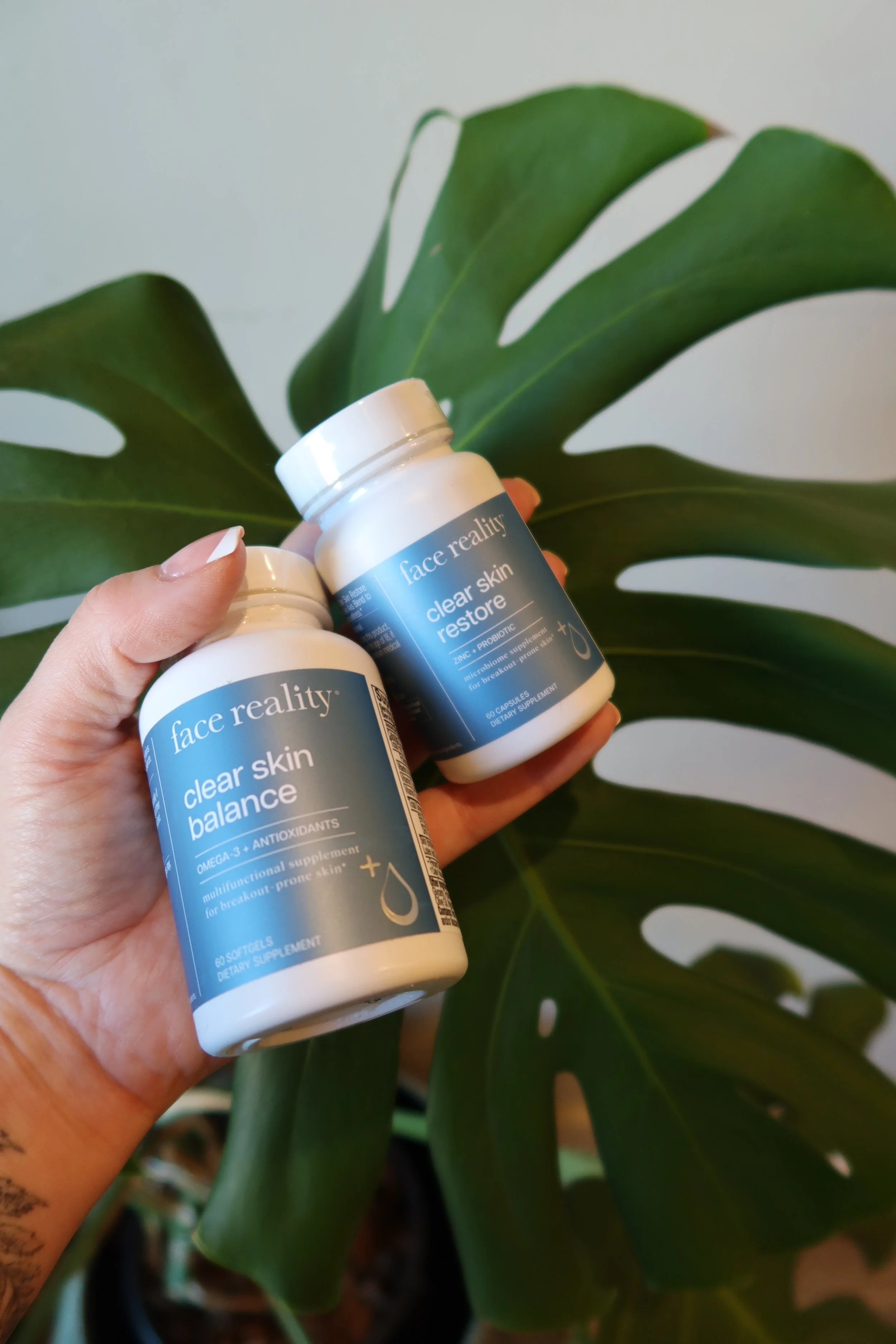 Two bottles of Face Reality dietary supplements, 'Clear Skin Balance' and 'Clear Skin Restore', held in front of a large green monstera leaf.