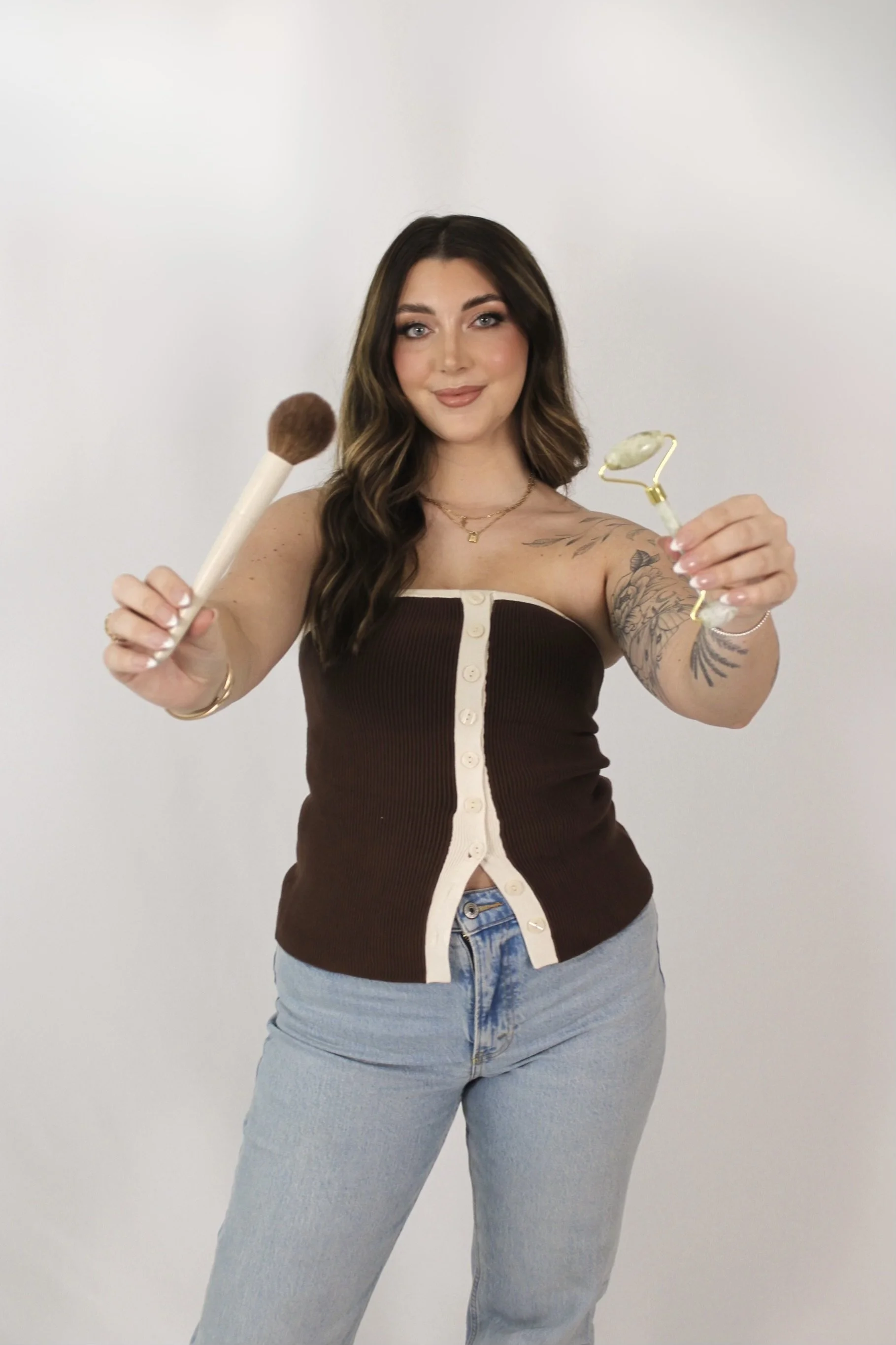 Woman with long, wavy brown hair holding a makeup brush in one hand and a jade roller in the other, smiling at the camera against a plain white background.