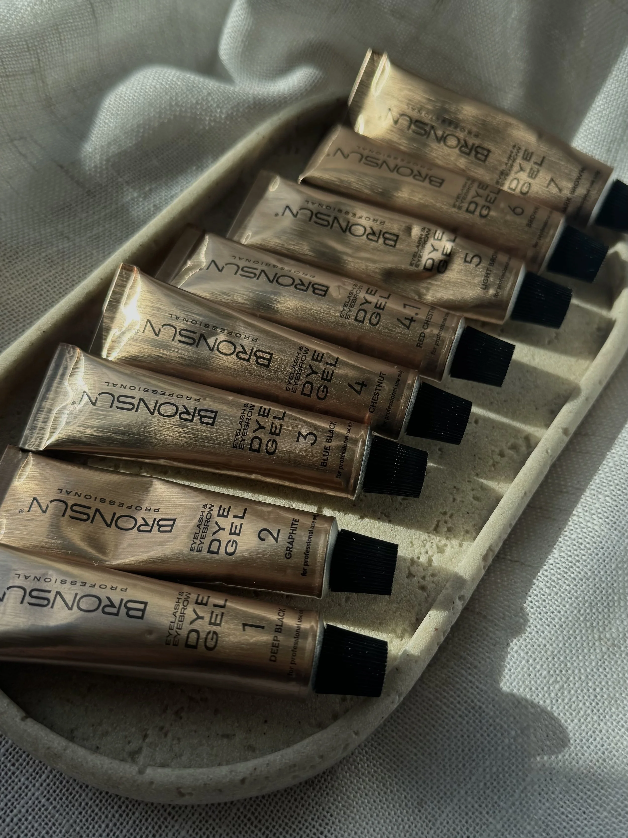 Six tubes of BROWSUN eyebrow dye gel arranged on a white, rectangular tray, with each tube labeled with color and number, including Deep Black, Graphite, Blue Black, Chestnut, Hazel, and Light Brown, on a white fabric surface.