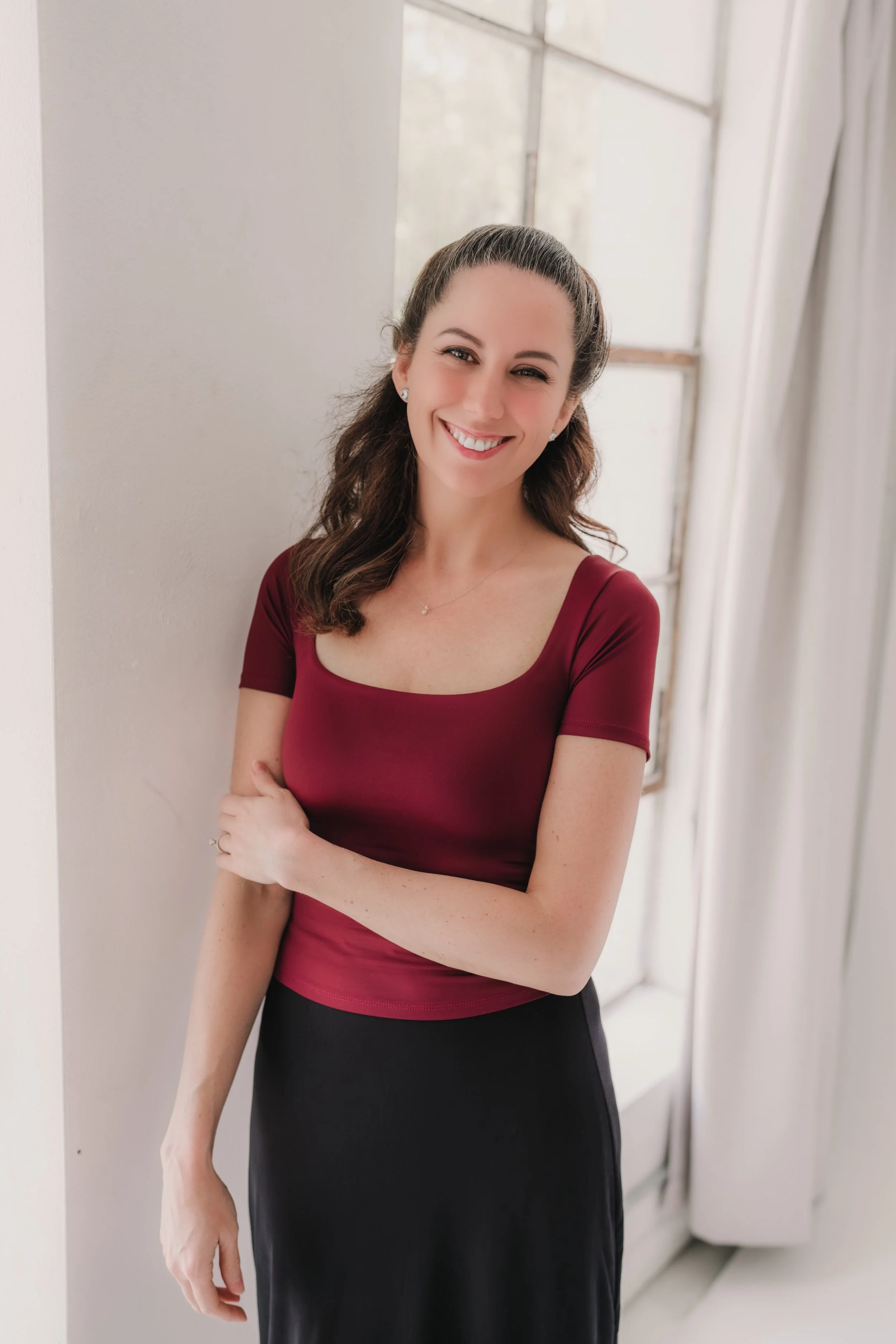 A smiling woman with long dark hair wearing sunglasses on her head, pearl earrings, and a light-colored collared shirt outdoors with green trees in the background.