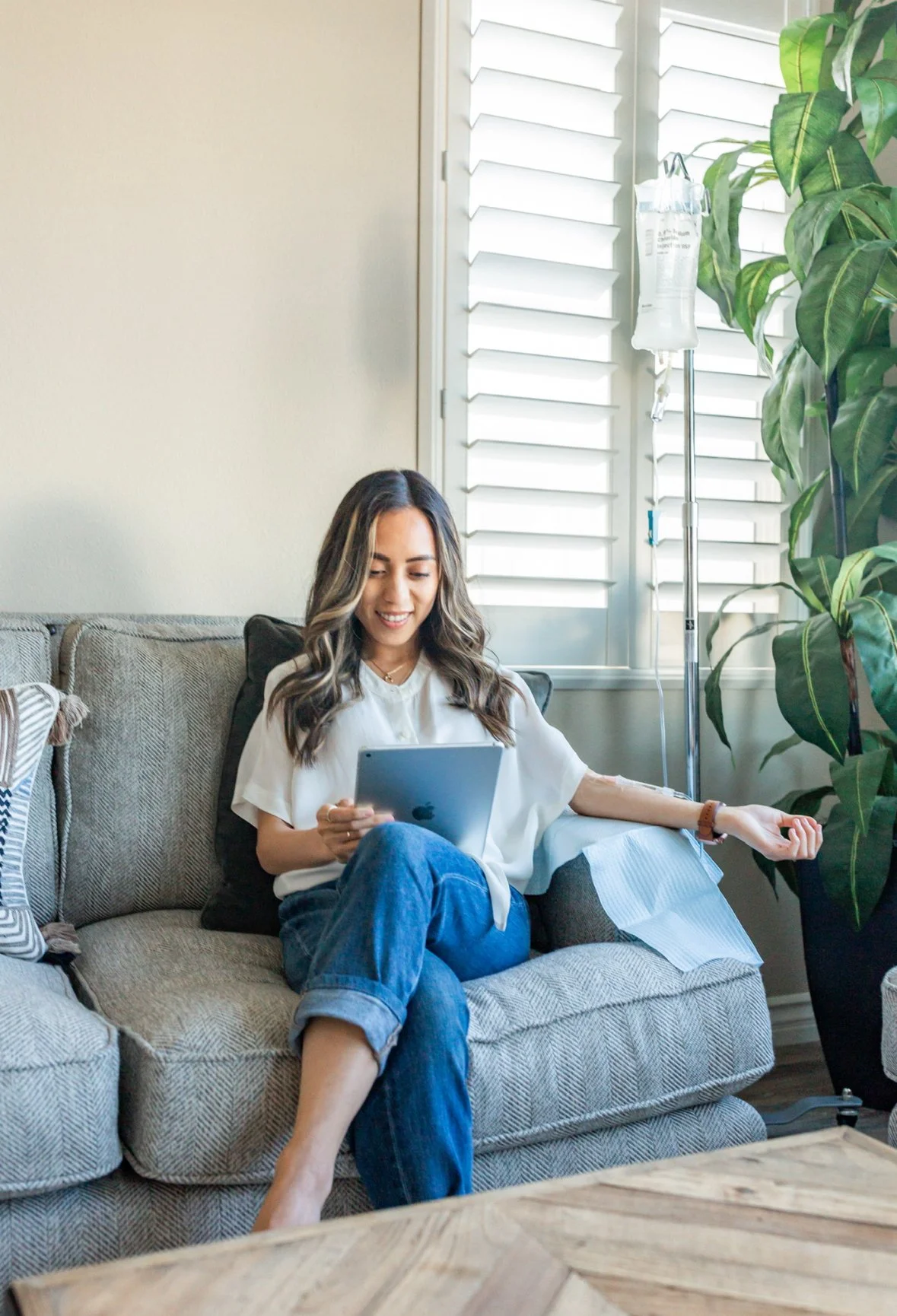 A young woman with long, wavy hair, wearing a white blouse and blue jeans, sitting on a gray couch with complimentary pillows. She is smiling and looking at a tablet. An IV drip stands beside her near a window with white plantation shutters, and a large green leafy plant is also nearby.