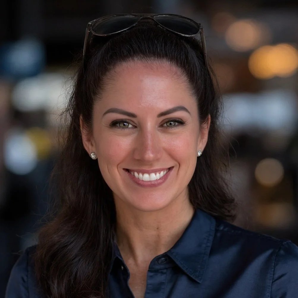A smiling woman with long dark hair wearing sunglasses on her head, pearl earrings, and a light-colored collared shirt outdoors with green trees in the background.