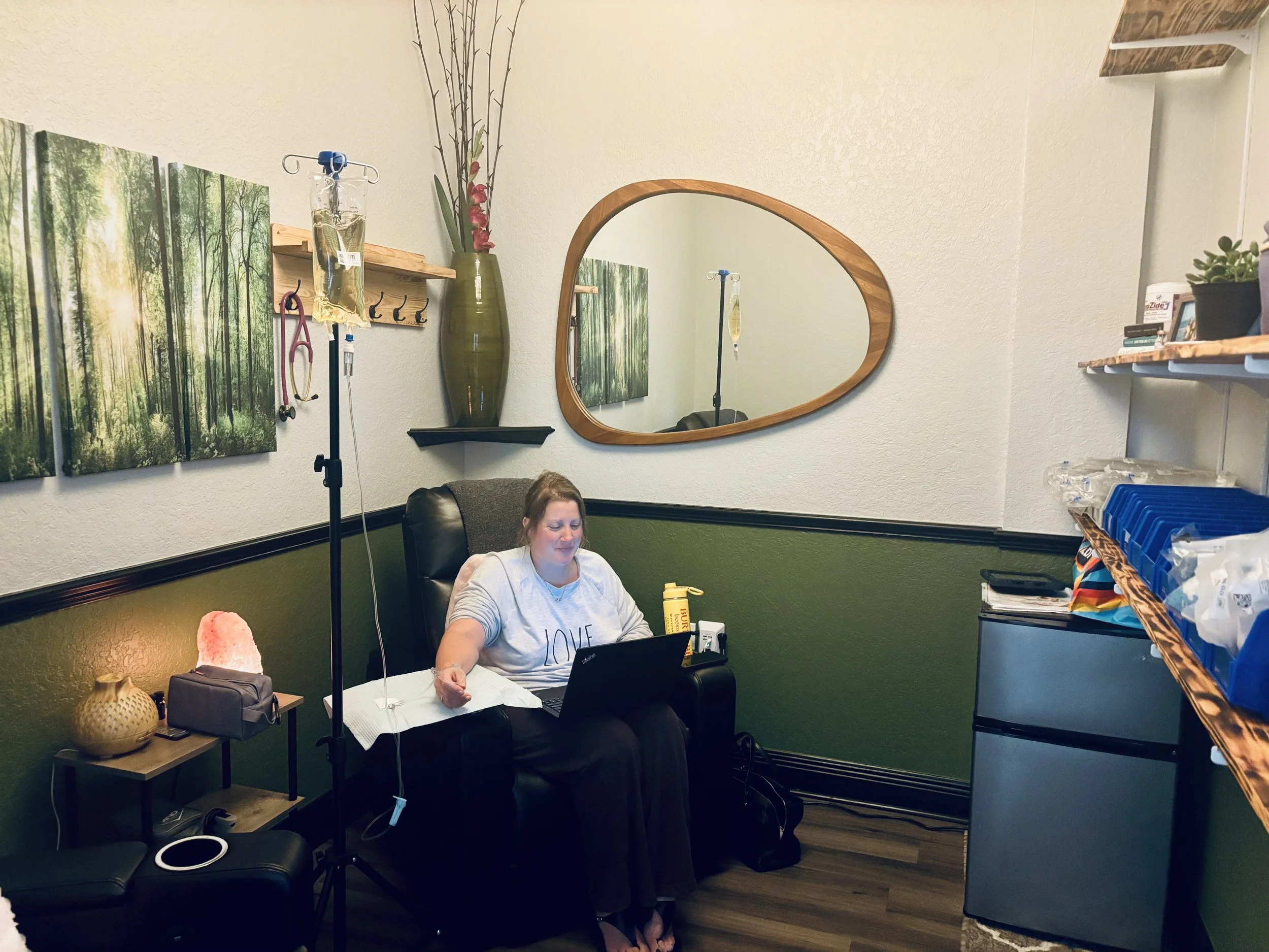 Woman in a hospital room sitting in a recliner chair with an IV pole nearby. The room has forest-themed artwork and a large, irregularly shaped wall mirror. There's medical equipment and supplies on shelves and a small table.