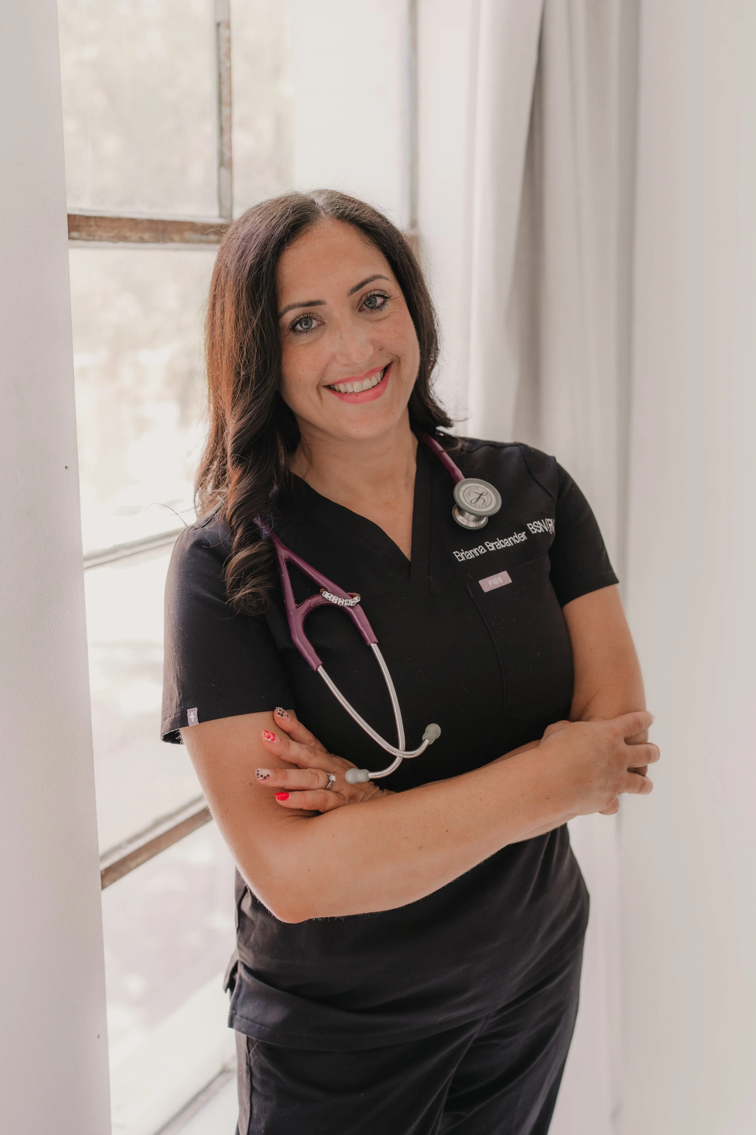 A woman with long brown hair, lipstick, and eye makeup smiling at the camera. She is wearing a red T-shirt with the United States Marine Corps emblem and a necklace with a small bird pendant. She is standing inside near a beige wall with a framed picture behind her.