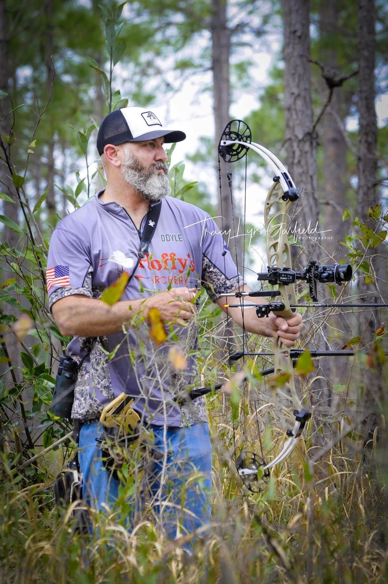 A man with a beard wearing a gray archery shirt and a cap, standing in a wooded area, aiming a compound bow.