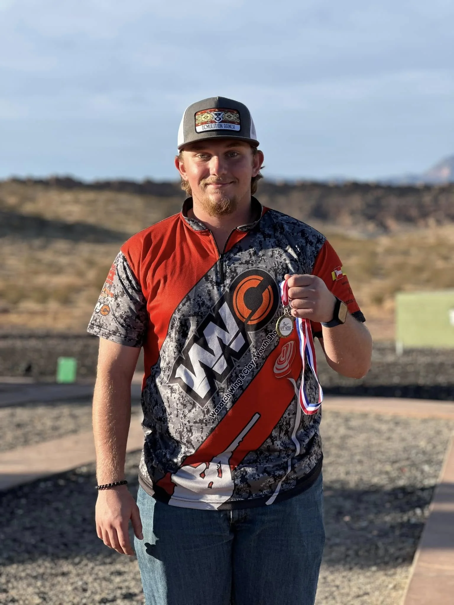 A young man wearing a red and black racing jersey and a baseball cap, holding a medal with a red, white, and blue ribbon outdoors in a desert landscape during daytime.
