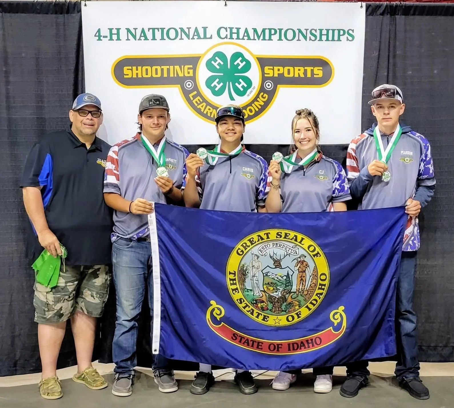 Group of five young athletes and one adult man standing, holding a flag of Idaho, at the 4-H National Championships in shooting sports. The athletes wear gray and blue uniforms with medals around their necks, and are smiling. The backdrop features a