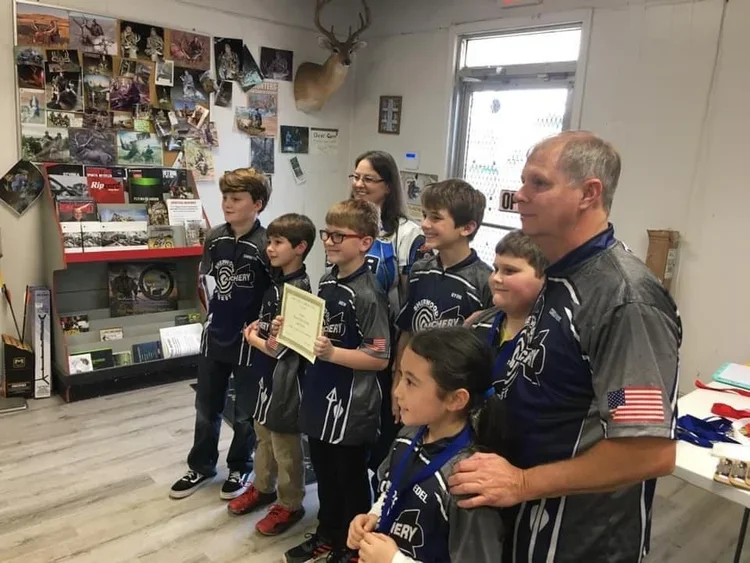 Group of children and an adult in a room decorated with photos and a mounted deer head, with one child holding a certificate.