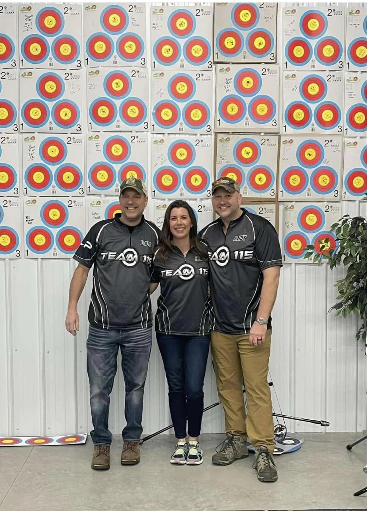 Three people standing in front of a wall of archery target posters, smiling and wearing matching black sports shirts.