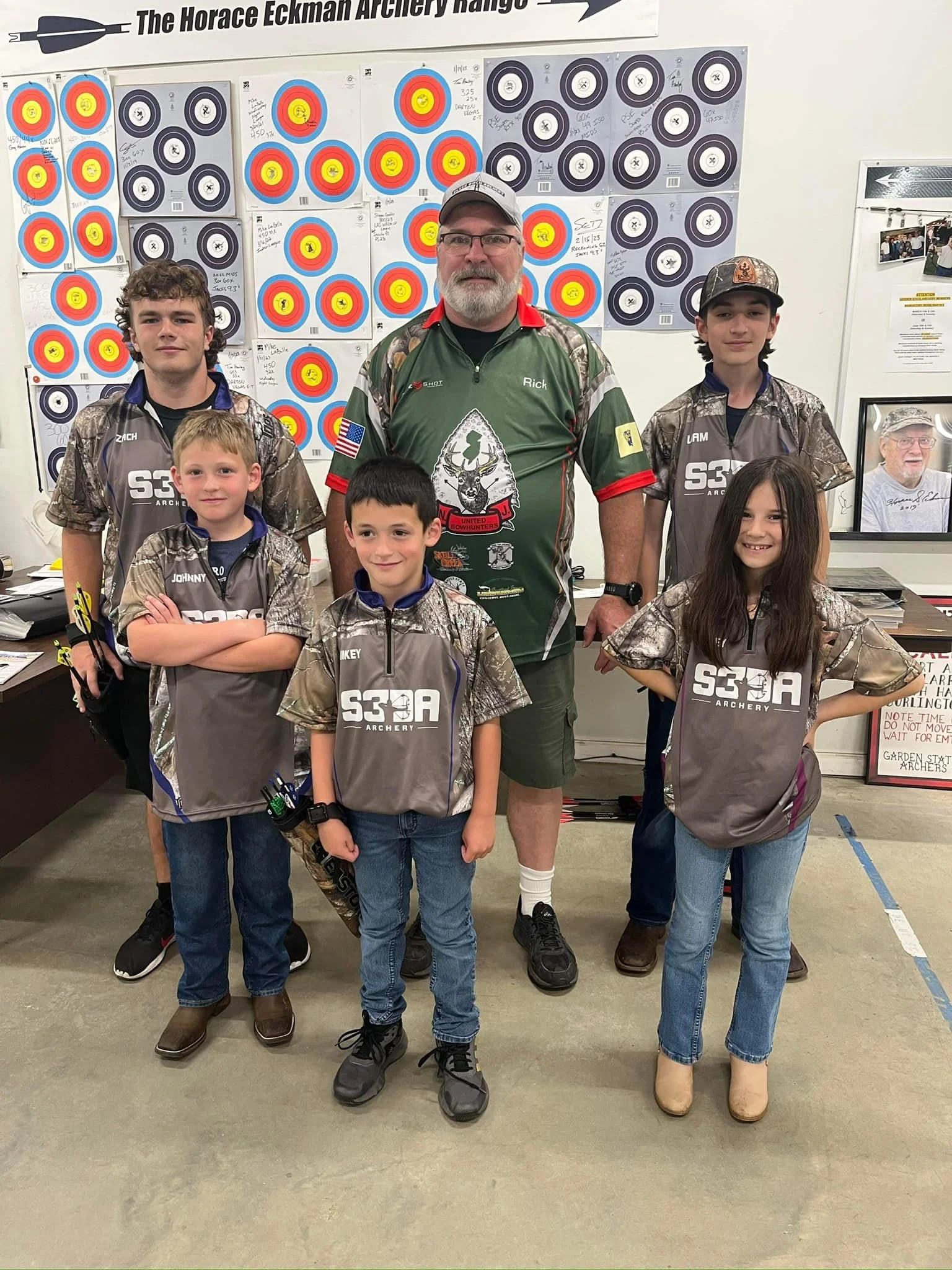 Group of six young archers, three boys and one girl, and their male coach standing in an indoor archery range in front of target papers with colorful bullseye targets. The coach is wearing a green and red sports shirt with the name 'Rick,' and the ch