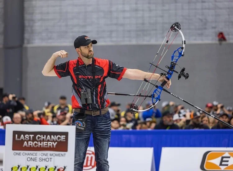An archer in red and black sportswear aiming a bow at an indoor archery competition, with a crowd in the background.