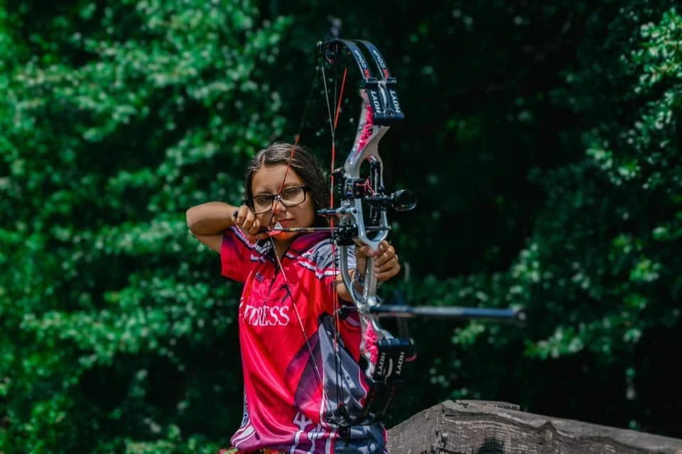 A young girl aiming a bow and arrow outdoors in a wooded area.