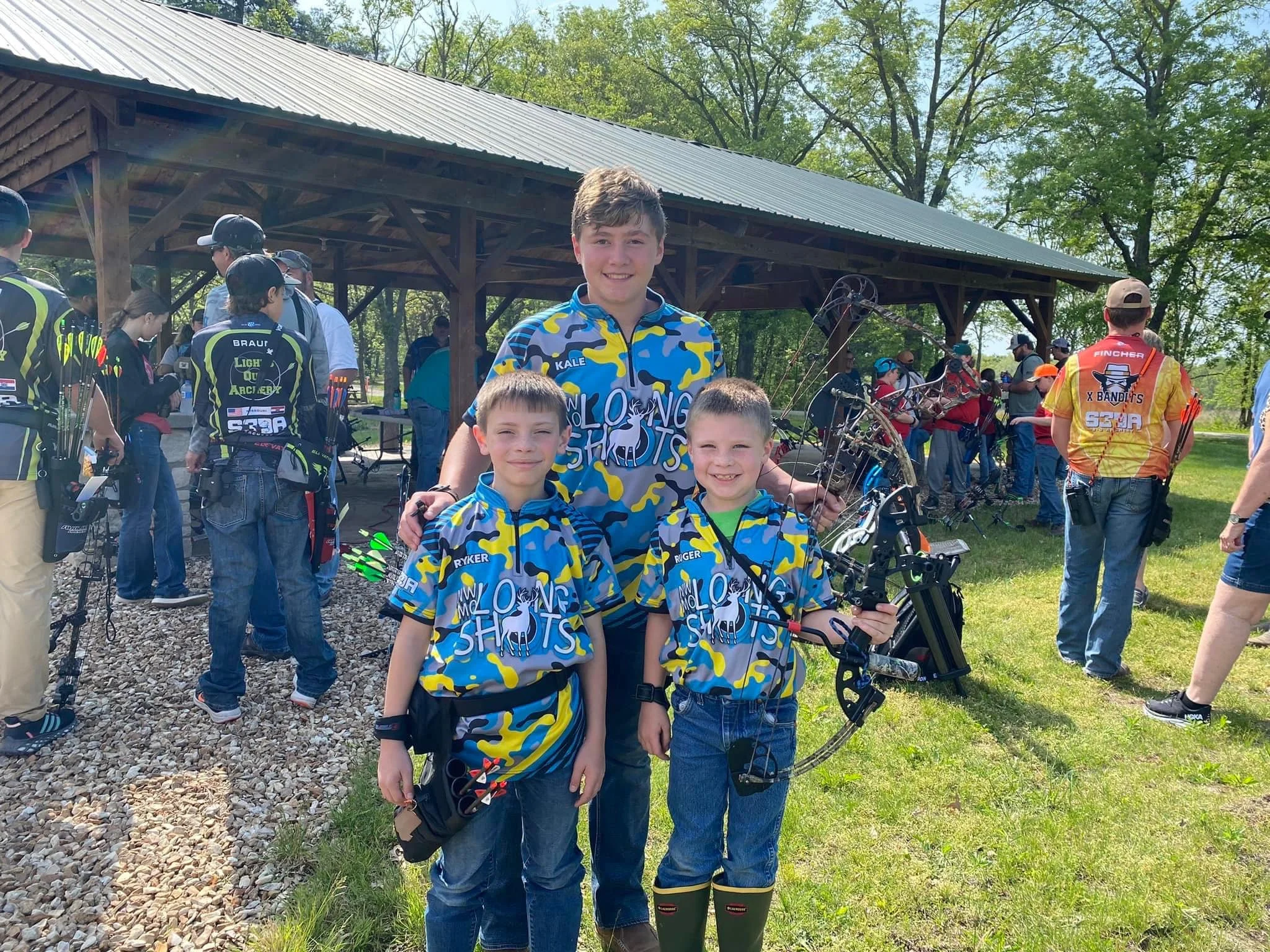 Four boys wearing matching blue, yellow, and gray camouflage shirts and holding archery equipment, smiling at the camera during an outdoor archery event, with other archers and a pavilion in the background.