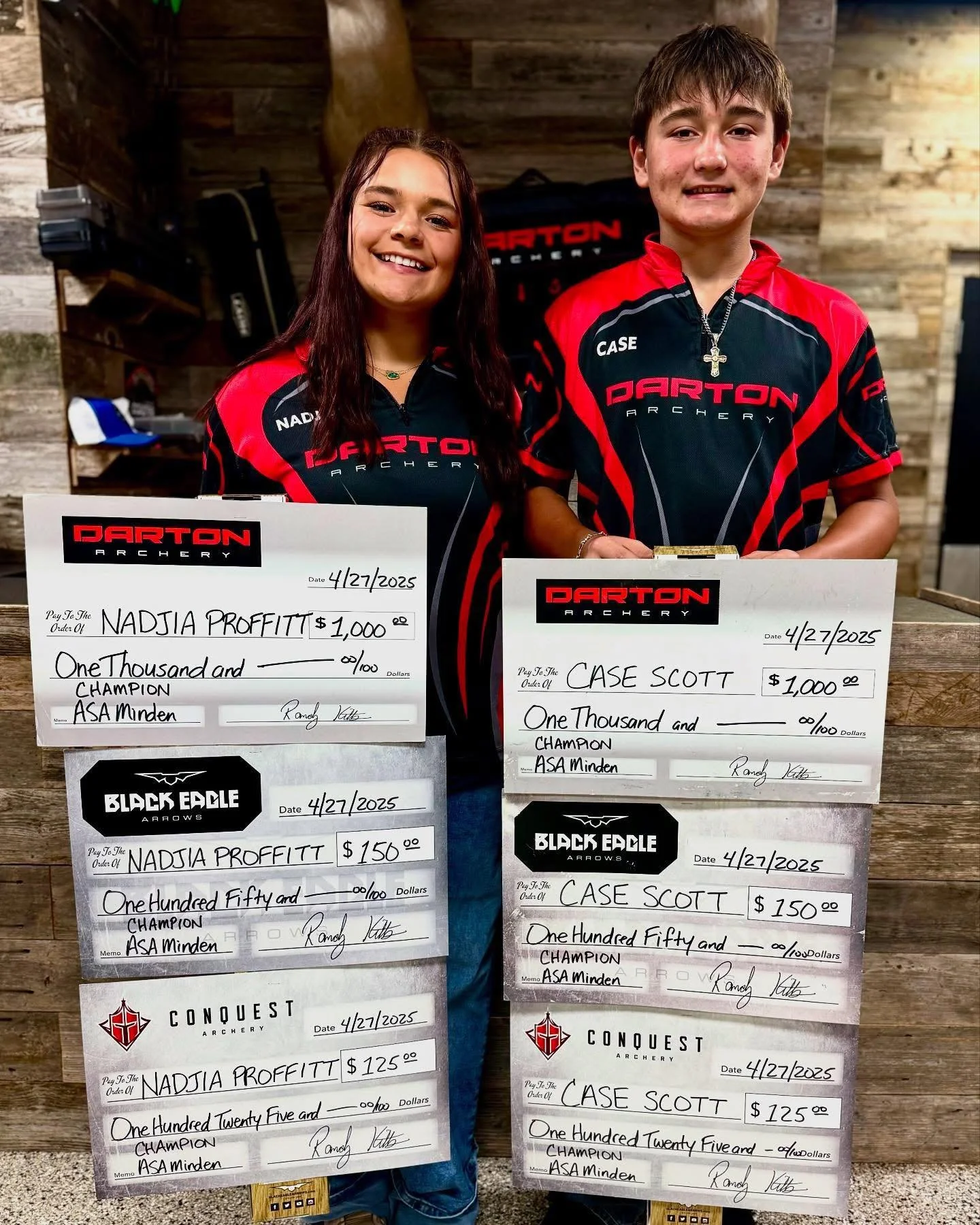 Two young archers wearing matching black and red Darton Archery shirts, standing indoors with wooden walls in the background. They are holding large checks for their archery achievements, dated April 27, 2025, awarded by Black Eagle Arrows and Conque
