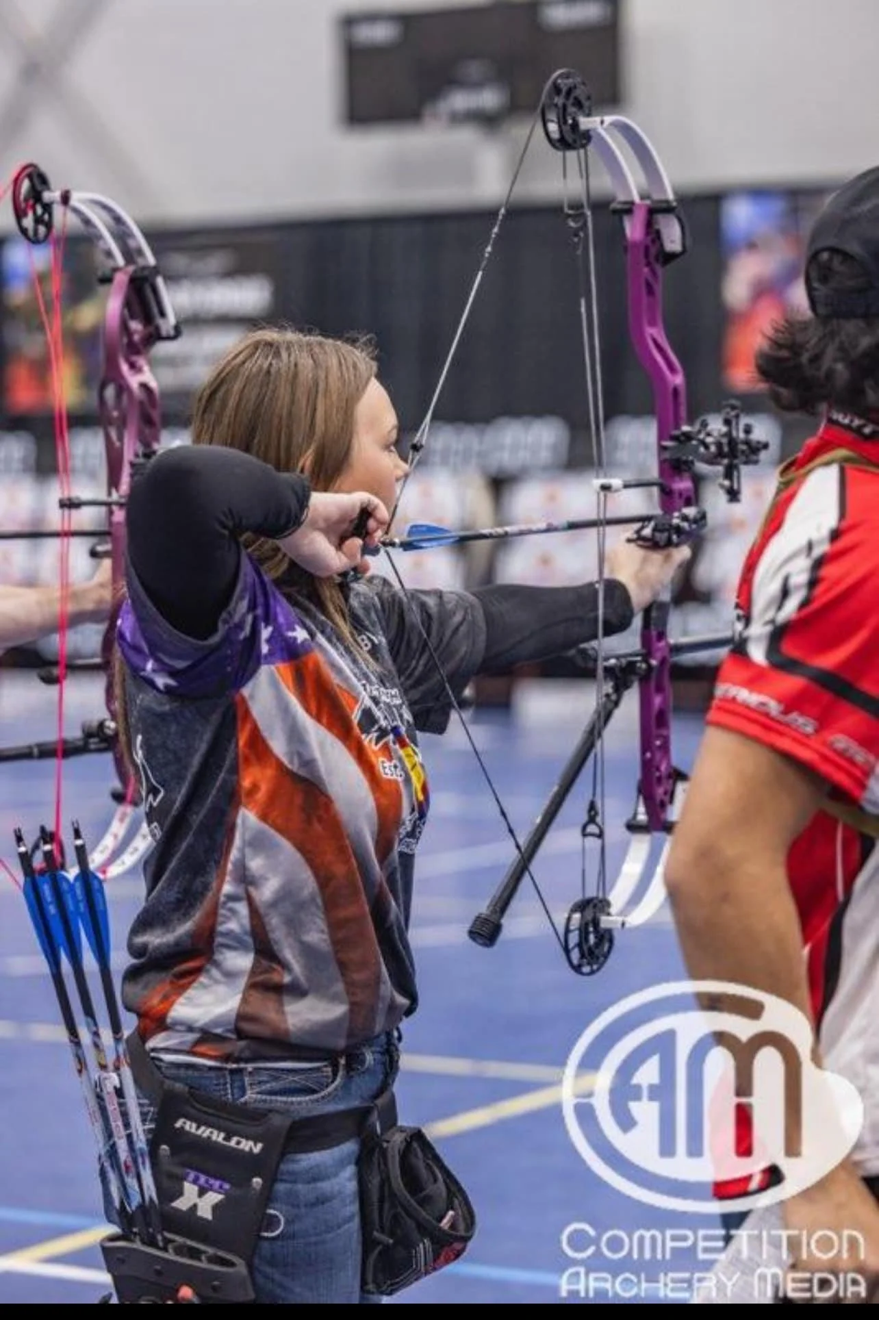 A female archer in a black, red, white, and purple uniform drawing a purple compound bow indoors, with arrows secured in a quiver on her hip, participating in an archery competition.