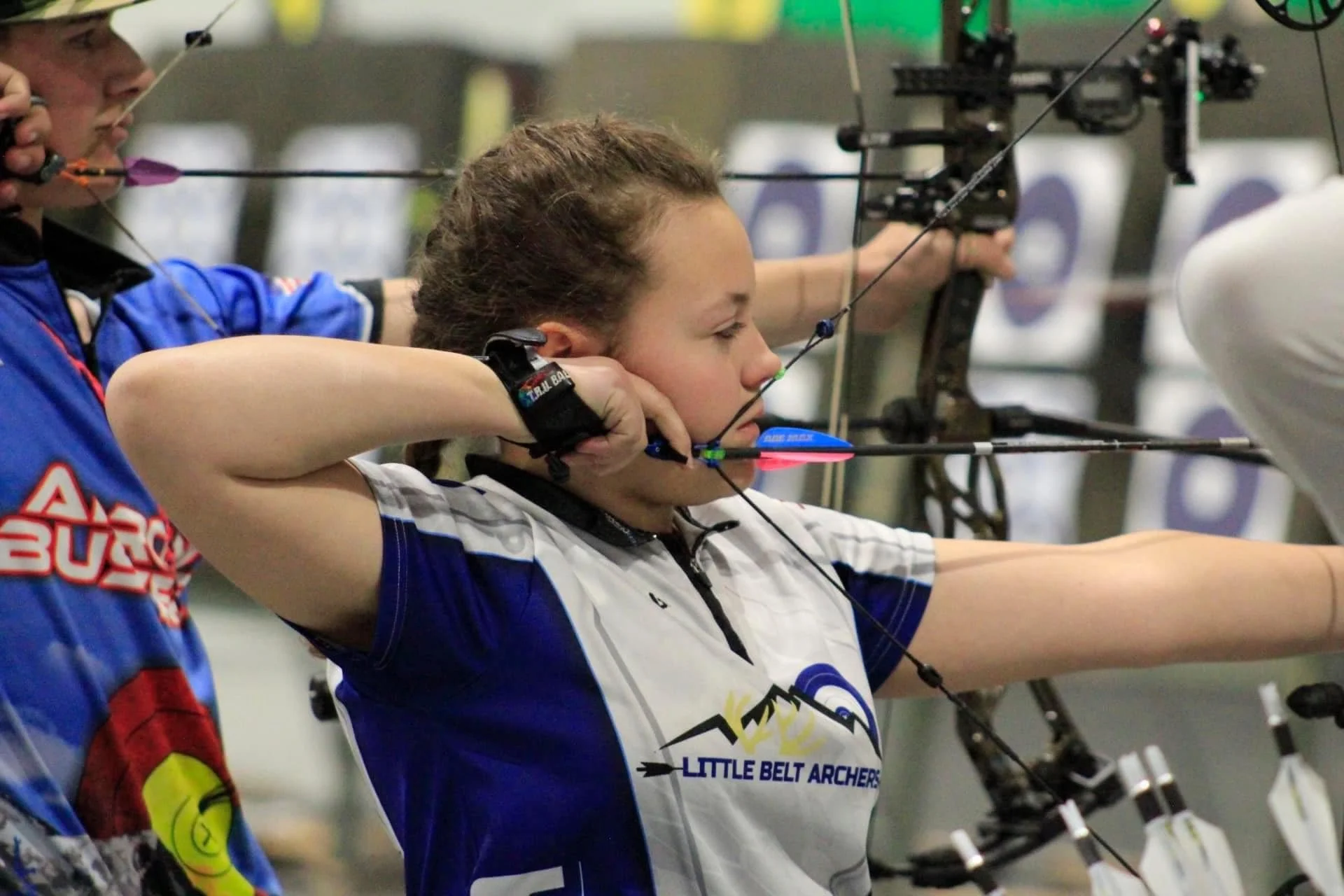 A female archer in a blue and white shirt drawing her bow during an indoor archery competition.