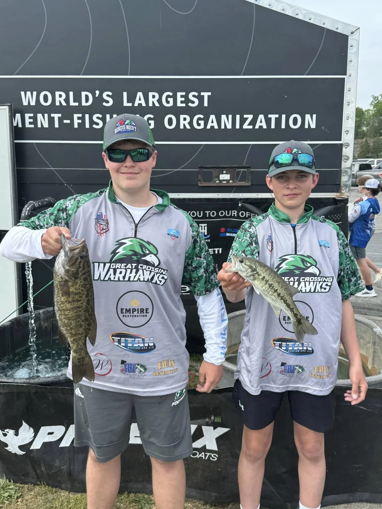 Two young male anglers holding fish in front of a black banner that reads 'World's Largest Ment-Fishing Organization,' wearing team jerseys with a logo 'Great Crossing Warhawks.'