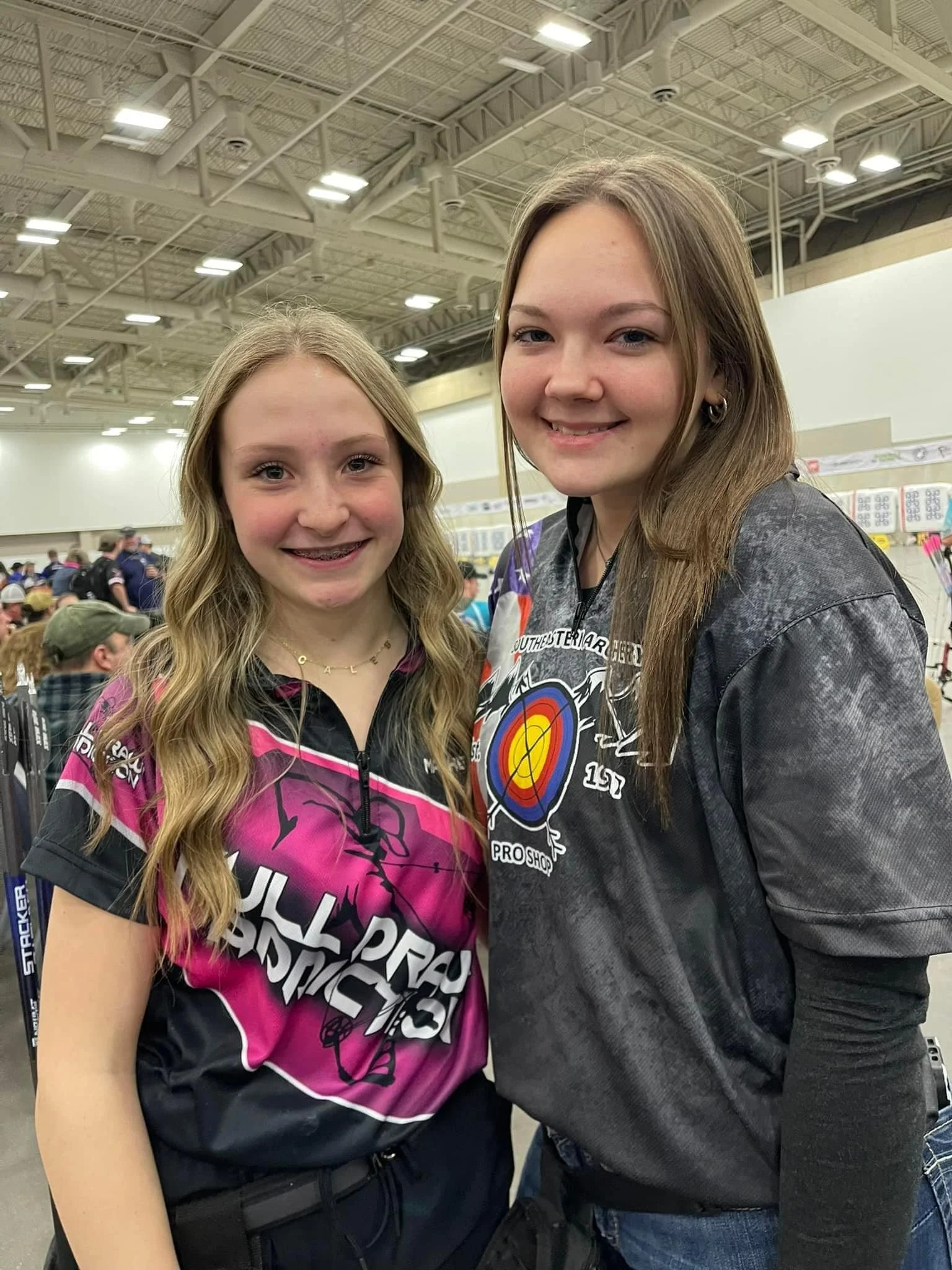 Two young girls smiling at an indoor event or convention, with numerous people and displays in the background.