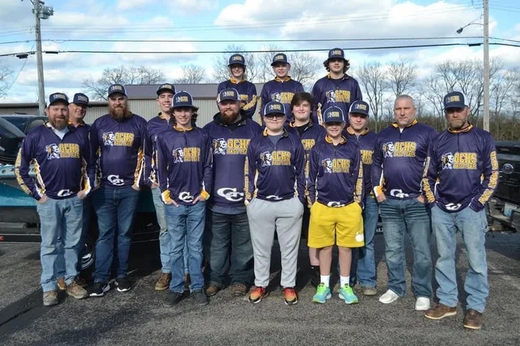 Group of people wearing matching purple and gold sports jerseys and hats, standing outdoors on a pavement with cars and trees in the background.