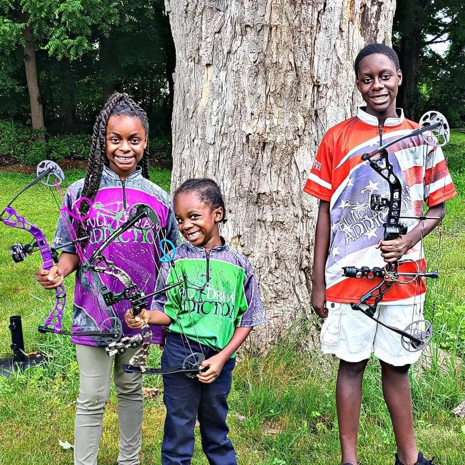 Three children smiling and holding bows and arrows outdoors near a tree in a grassy area.