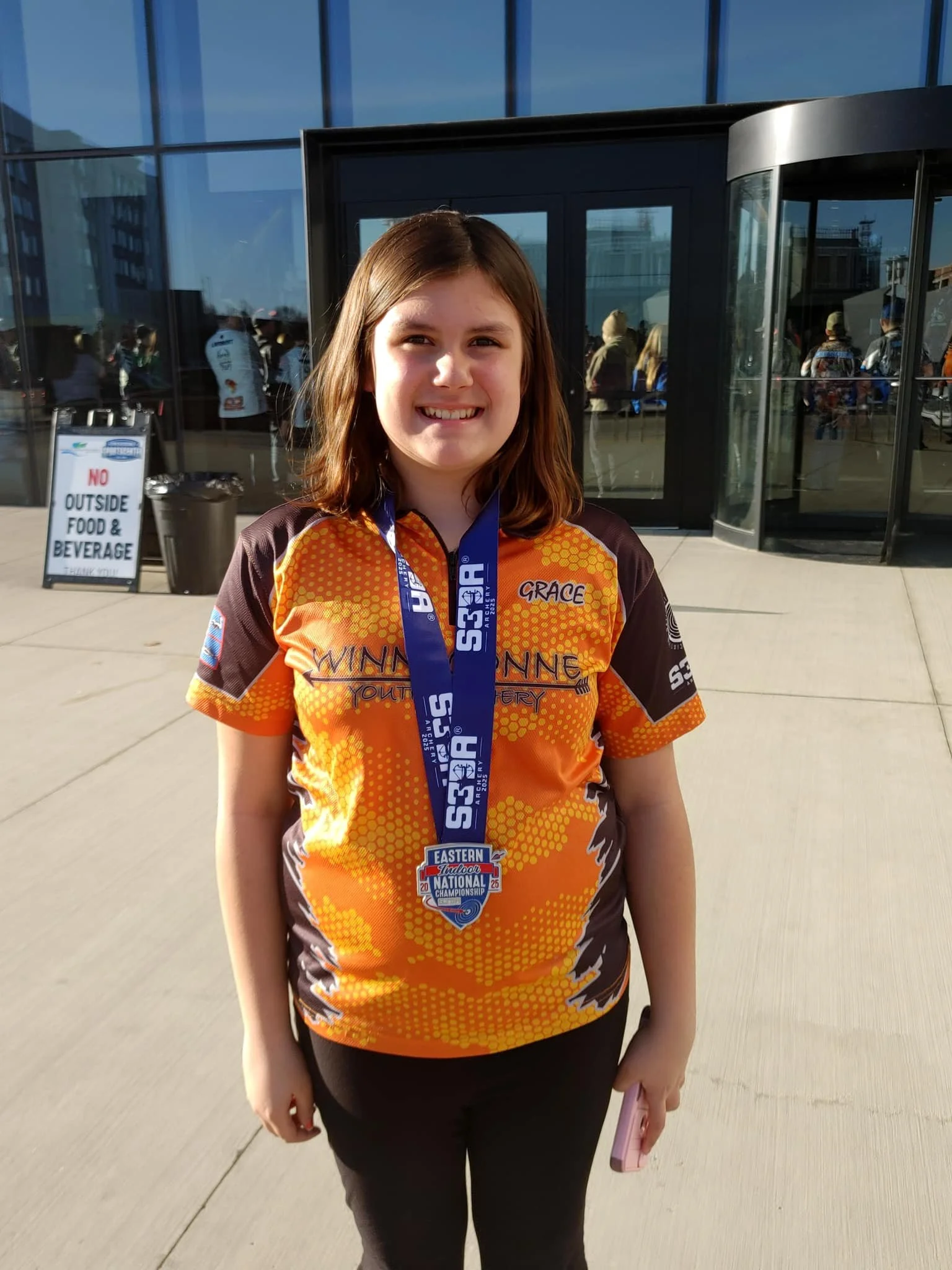 Young girl wearing an orange and brown sports jersey with a medal around her neck, standing outside a building, smiling at the camera.