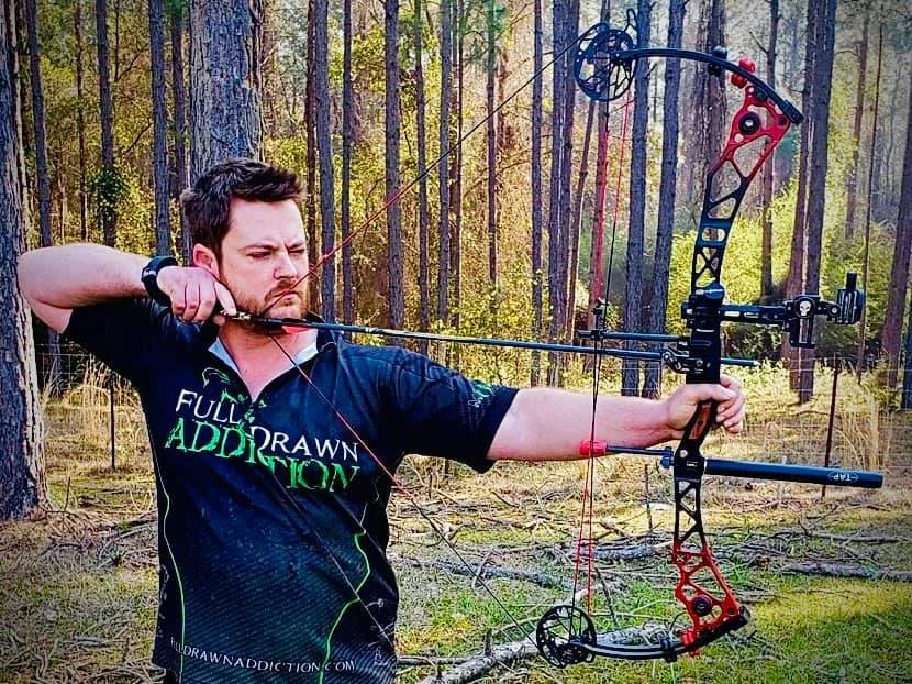 Man with dark hair and beard aiming a compound bow in a forest setting, wearing a black shirt with green text.