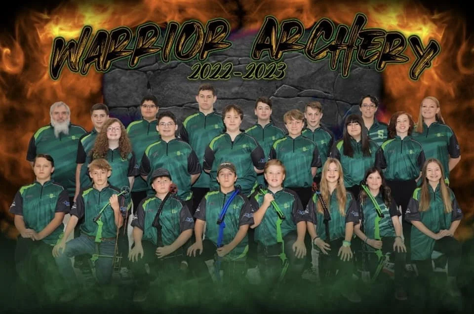 A group of young archers and their coach posing for a team photo, wearing matching green and black uniforms, with a background of flames and a rock wall, and the text 'Warrior Archery 2022-2023' at the top.