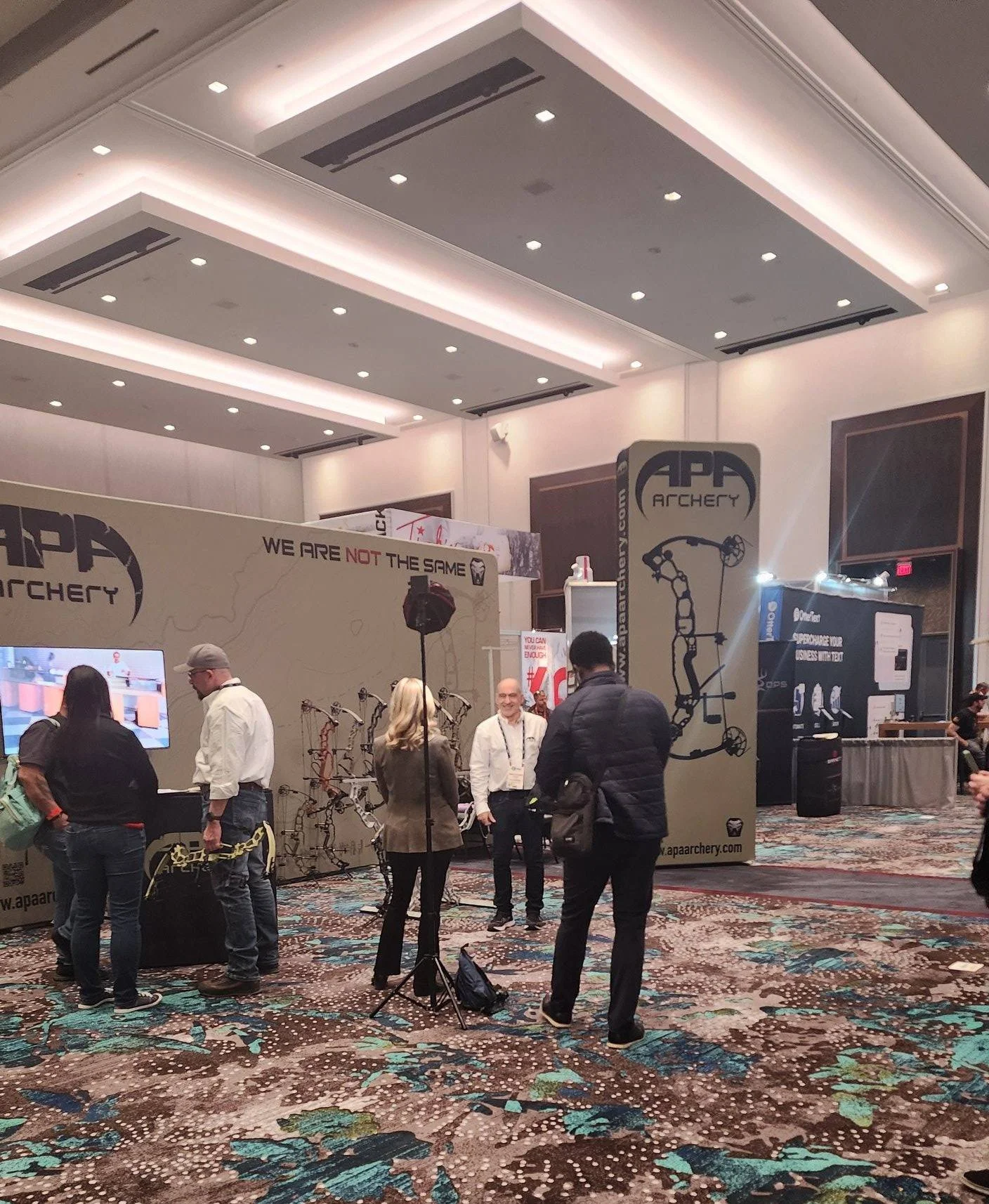 A trade show booth for APA Archery with people interacting, displaying bows and archery equipment, decorated with a large map and the message 'We Are Not The Same'