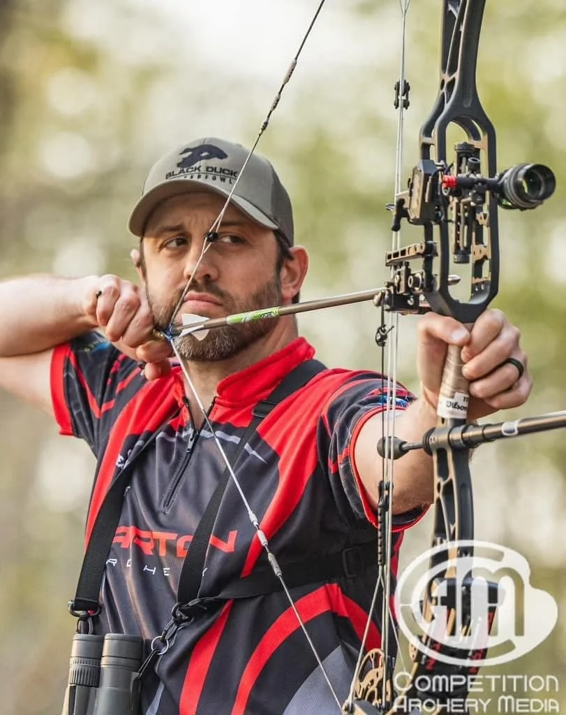 Man aiming a compound bow with a scope outdoors, wearing a baseball cap and a red and black sports shirt.