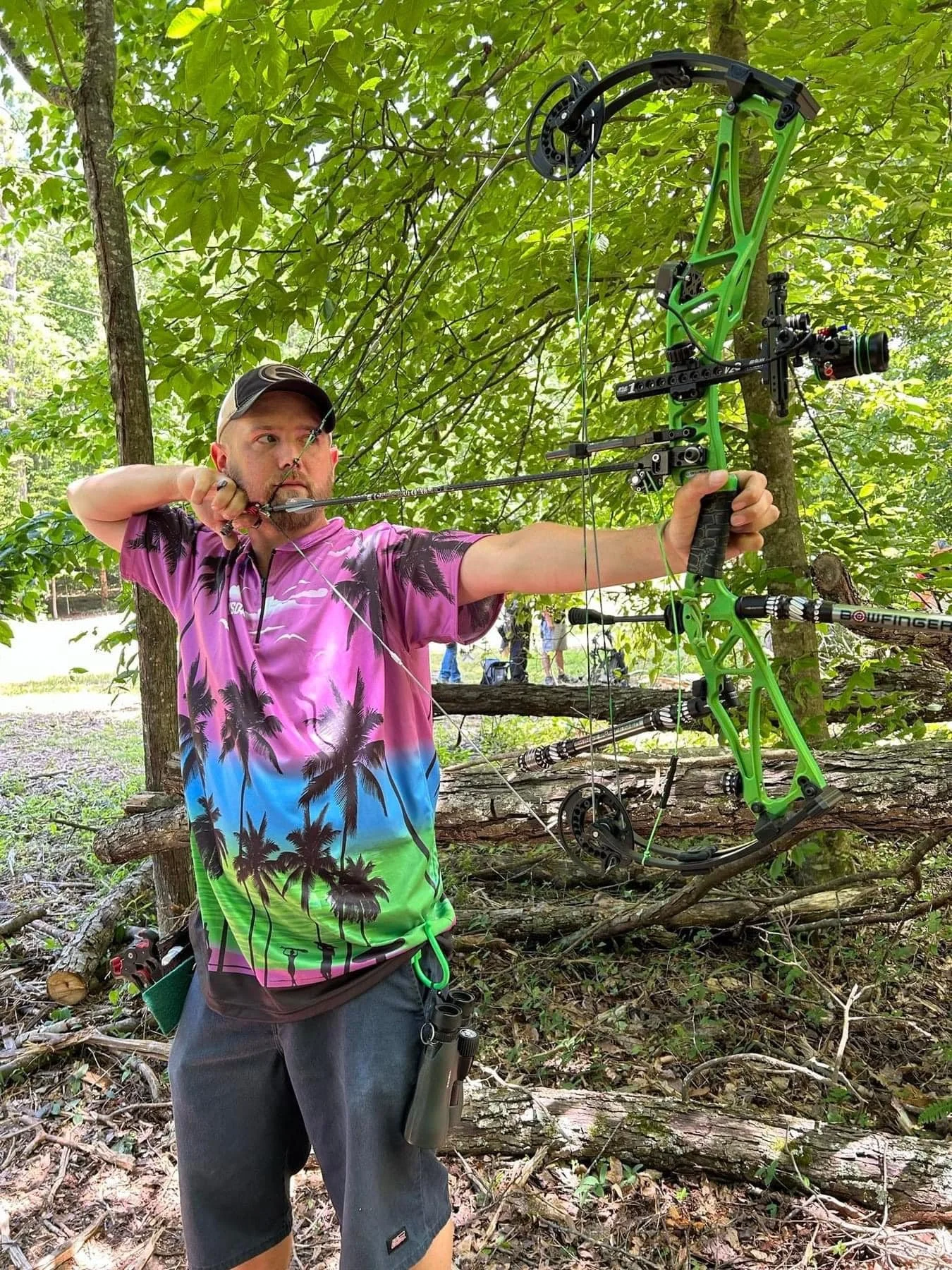 A man aiming a green compound bow with a black scope in a wooded area, wearing a colorful tropical shirt and a cap.