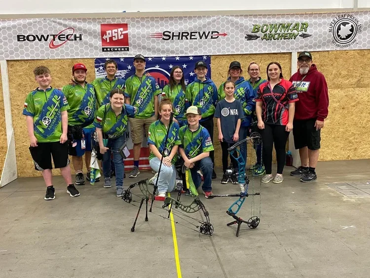 Group of young archers in green shirts posing indoors with bows, surrounded by archery banners and logos.
