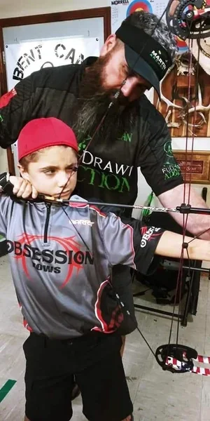A bearded man wearing a black headband and black sports shirt assisting a young girl with a bow and arrow at an indoor archery range.