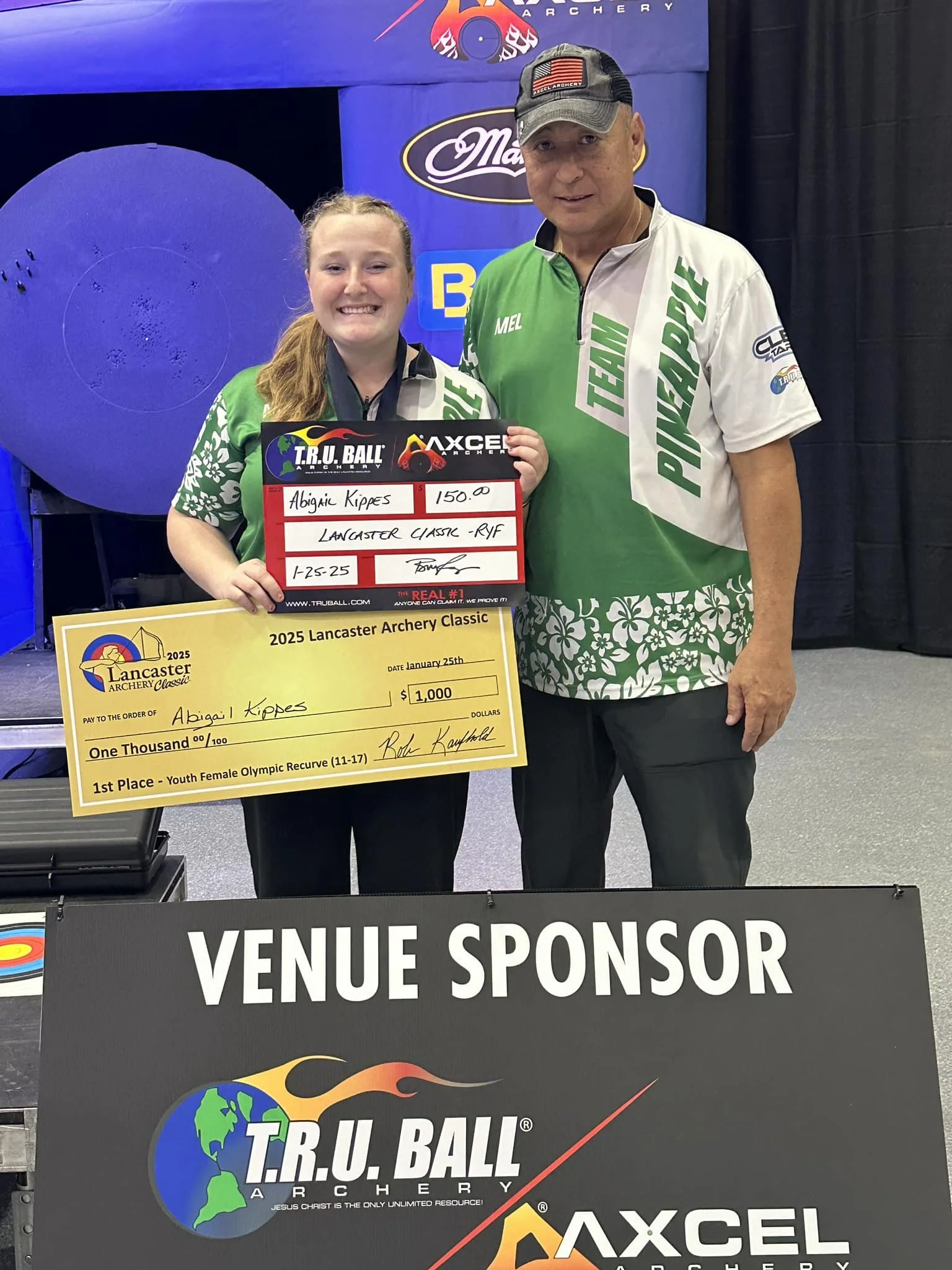 A young girl and a man standing together at an archery event. The girl is holding a large check for $1,000 and a plaque indicating first place in the Youth Female Olympic Recurve category. Both are wearing green and white team shirts, and the girl is