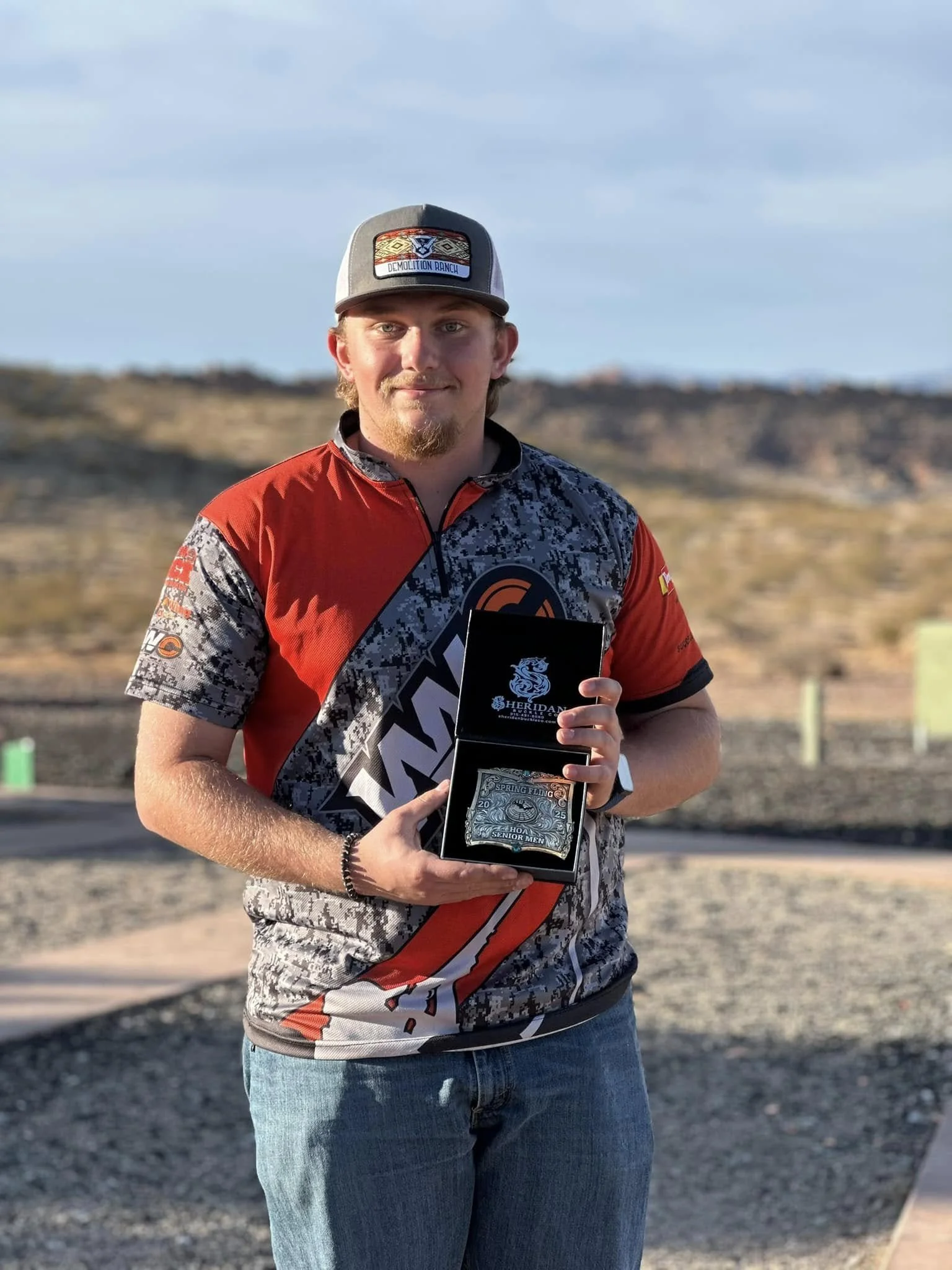 A young man with a beard and mustache in casual clothes holding a black box with a certificate or award inside, outdoors in a desert-like area during daytime.