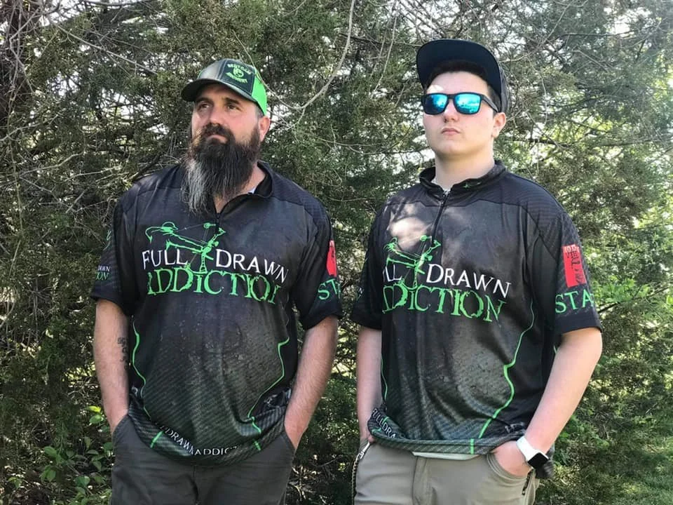 Two men standing outdoors in front of trees, wearing matching black sports jerseys with green and white text that says 'Full Drawn Addiction' and a graphic of a person drawing a bow. One man has a beard, wearing a green and black cap, and has tattoos