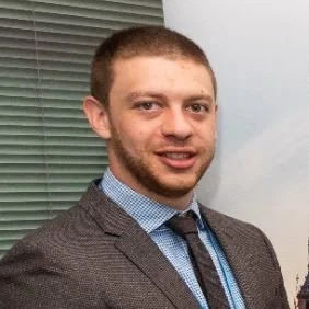 A young man with short hair and a beard, wearing a brown blazer and blue shirt, smiling in front of a window with closed blinds.