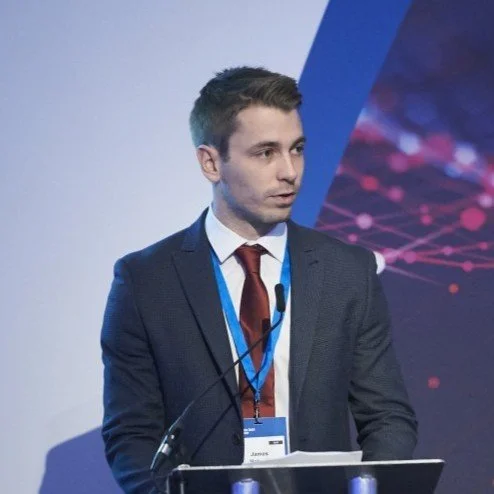 A young man in a suit speaking at a conference or presentation, with a microphone and a lanyard around his neck.