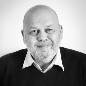 A black-and-white portrait of a middle-aged man with a beard, smiling, wearing a collared shirt and a dark sweater.